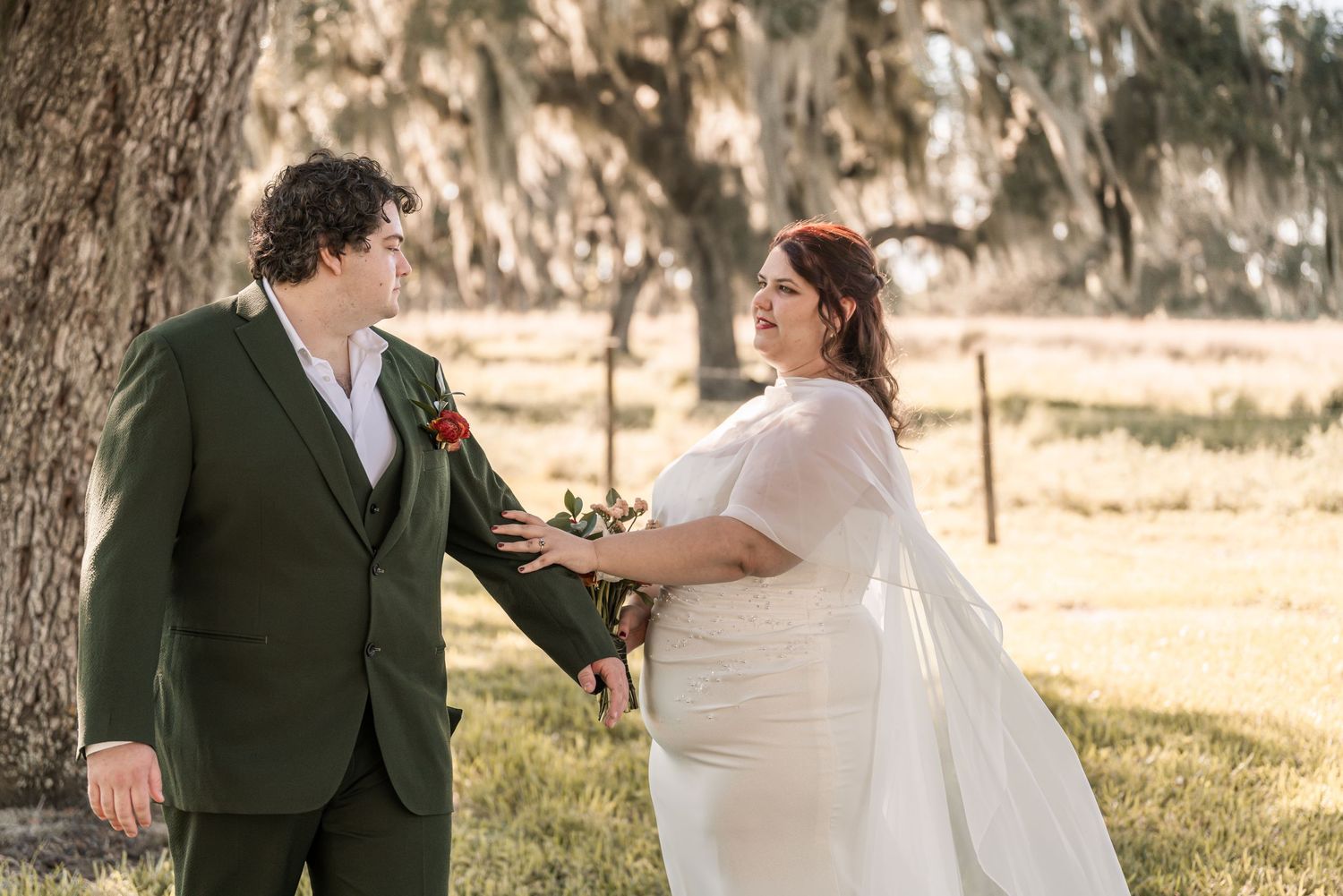 A romantic wedding couple shares intimate moments in a sun-drenched field with Spanish moss draped trees in the background.
