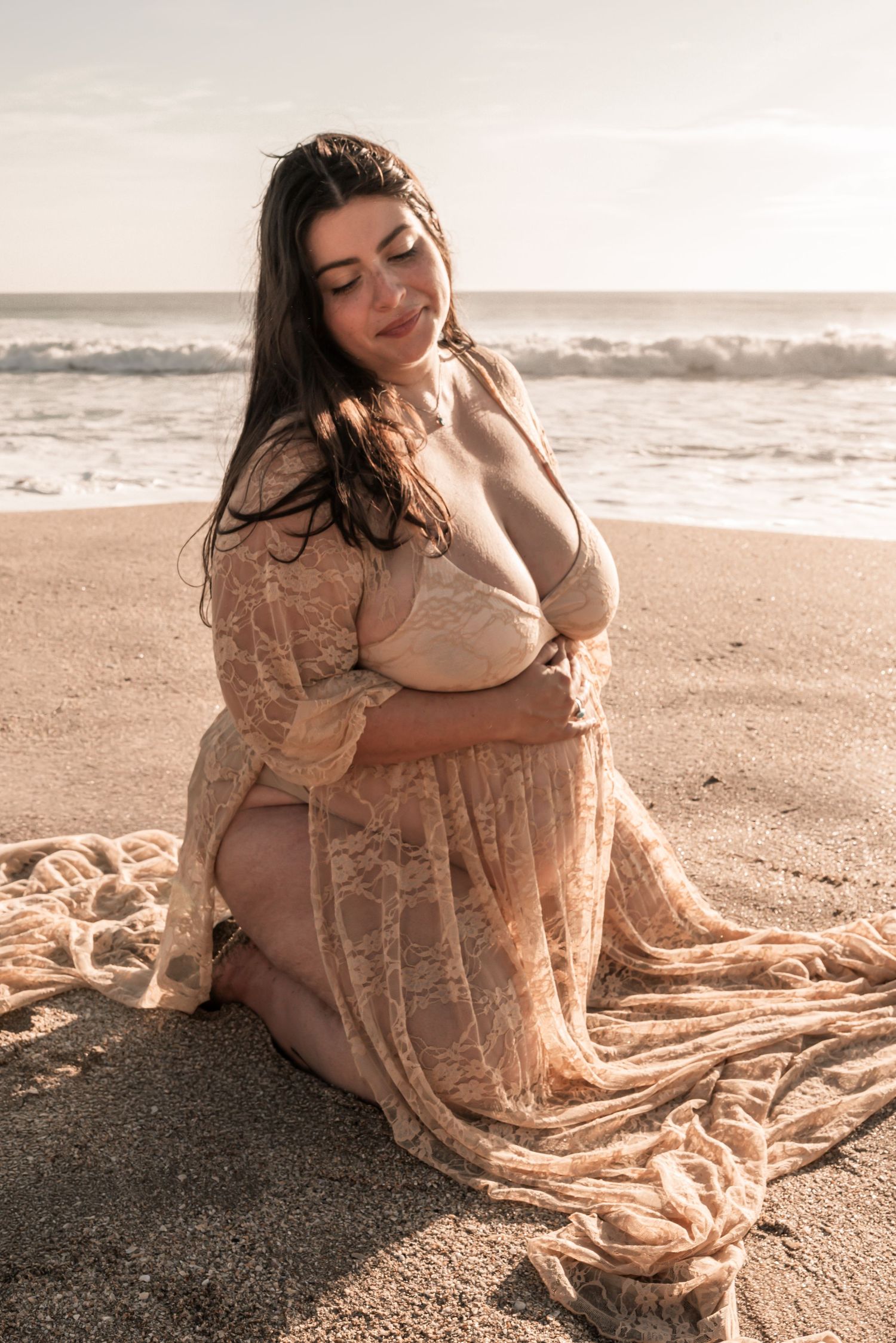Pregnant woman in flowing beige lace dress poses gracefully on sandy beach at sunset.
