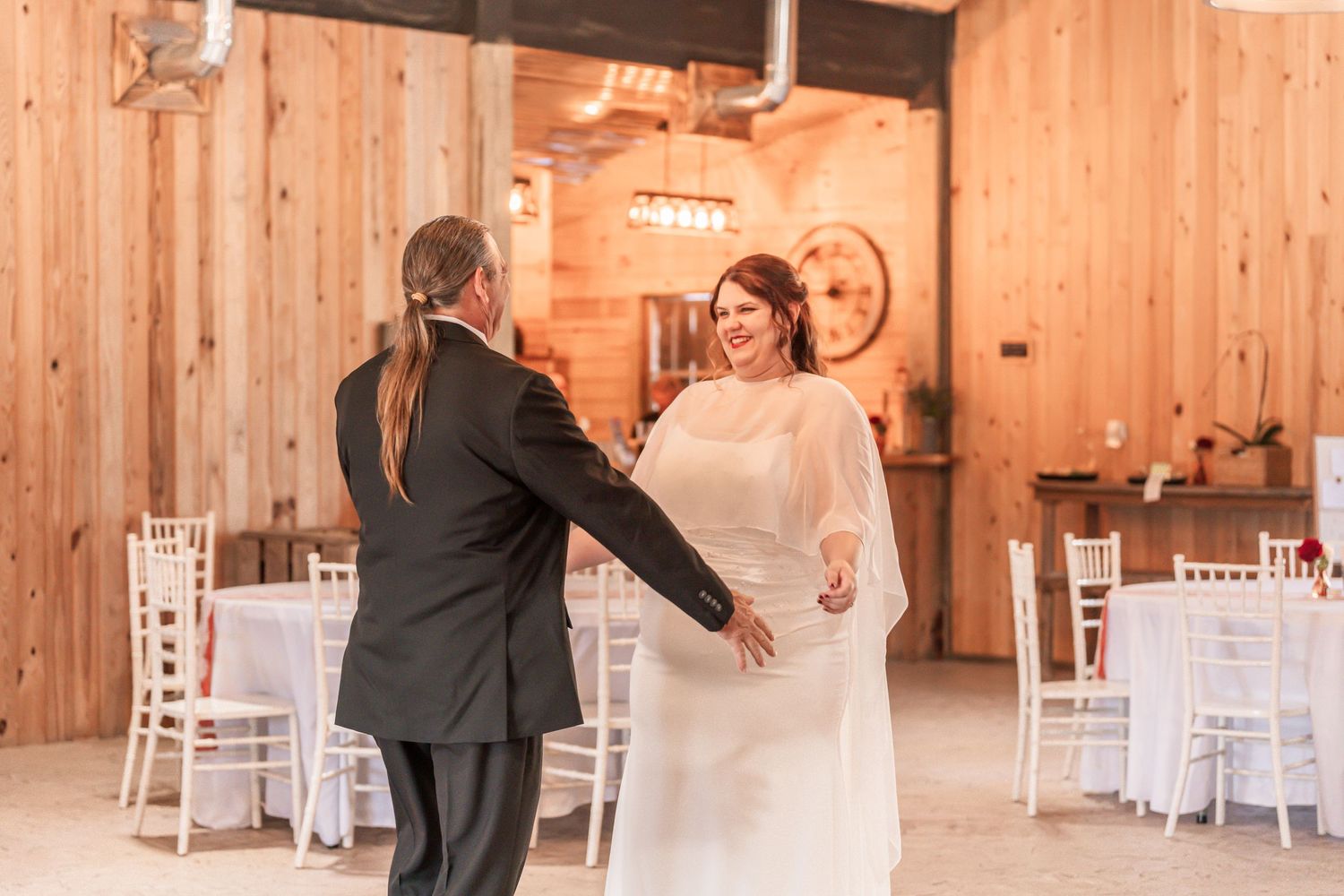 A couple shares their first dance in a rustic wooden barn venue decorated with white tables and chairs.