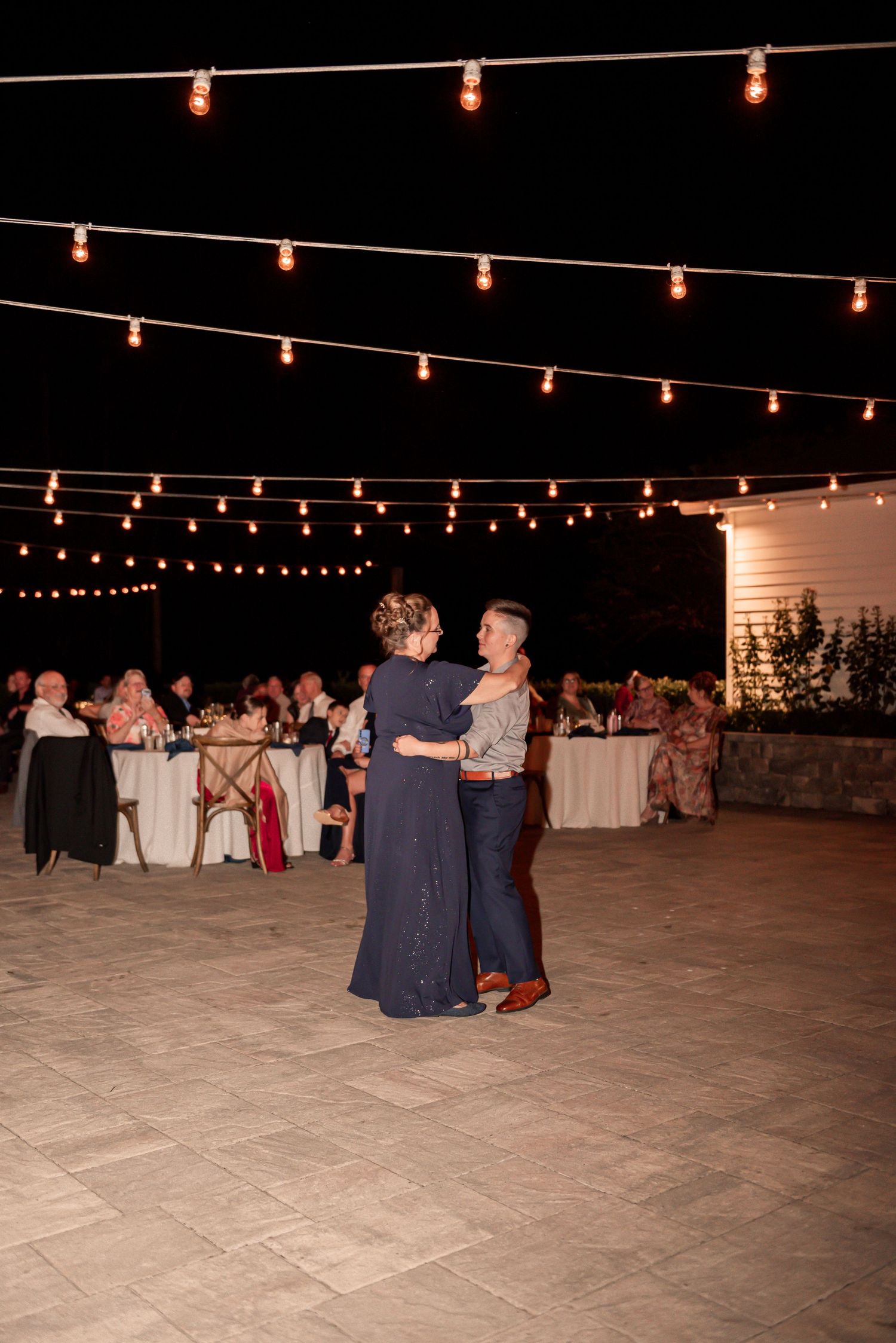 String lights illuminate an outdoor dance floor where couples dance at a nighttime wedding reception.