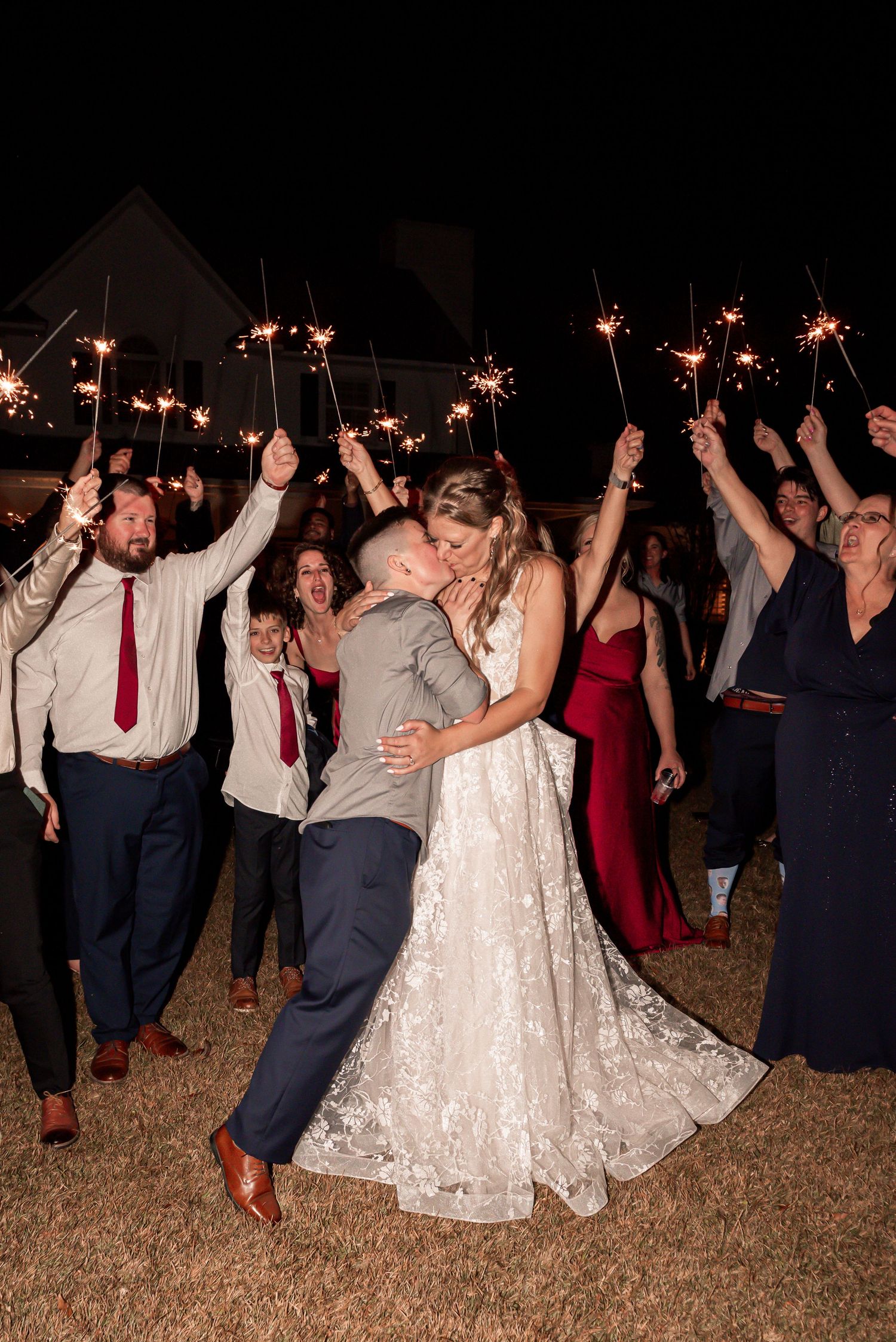 Wedding party celebrates on lawn at night holding sparklers while dancing and having fun at outdoor reception.