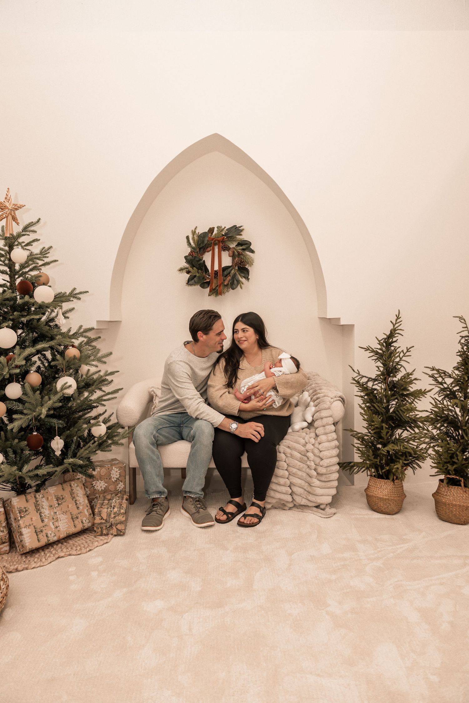 Cozy winter family photoshoot setup with evergreen trees and white archway against neutral backdrop.