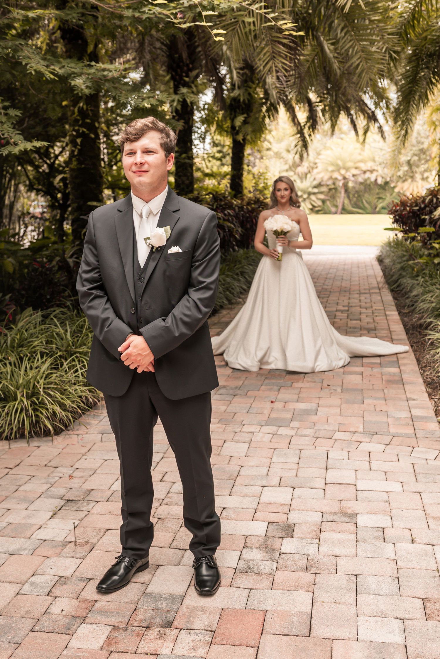 Groom in black tuxedo stands on brick path as bride in white gown approaches in lush garden for first look.