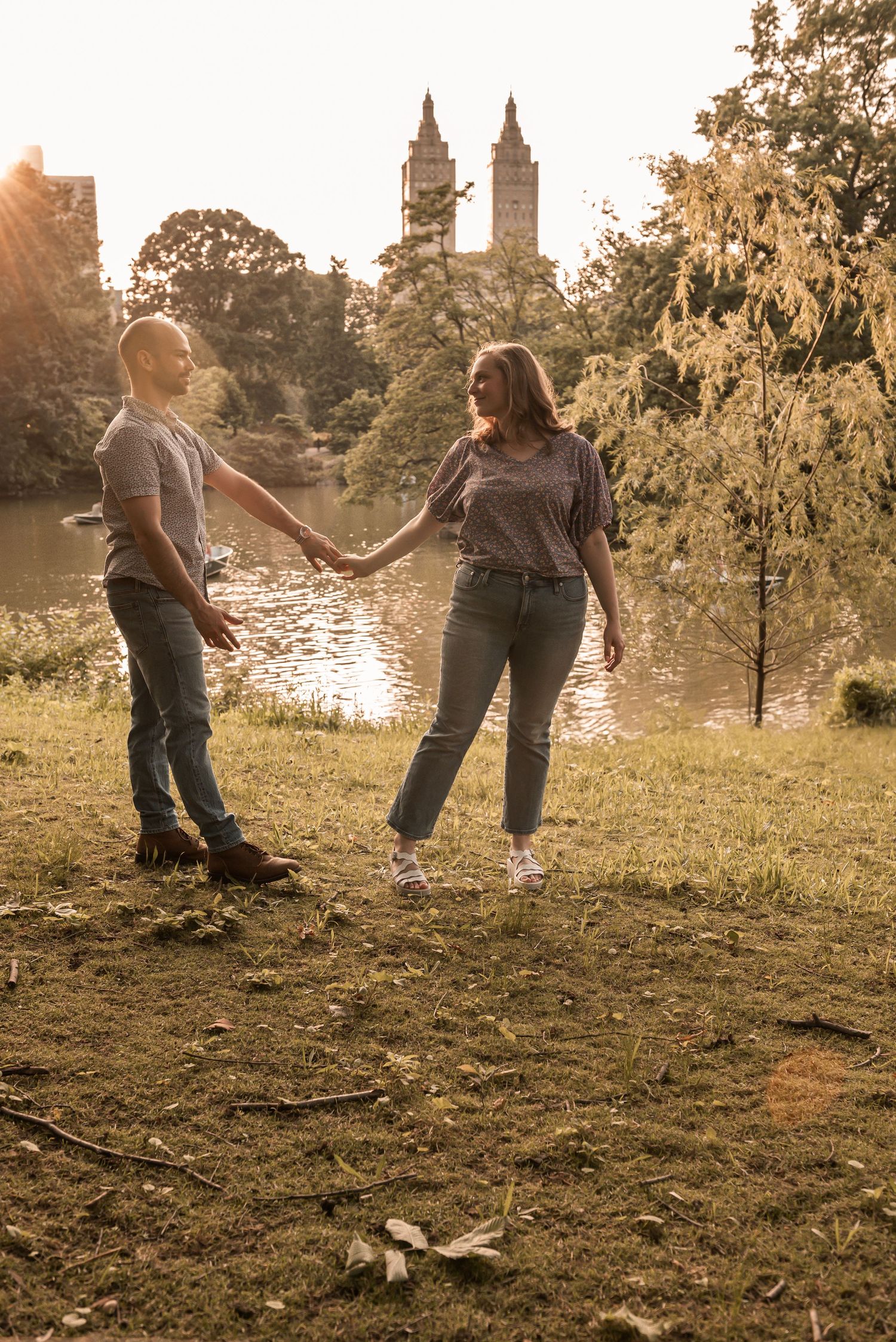 A couple holds hands at sunset in Central Park with iconic Manhattan buildings visible in the golden hour light.