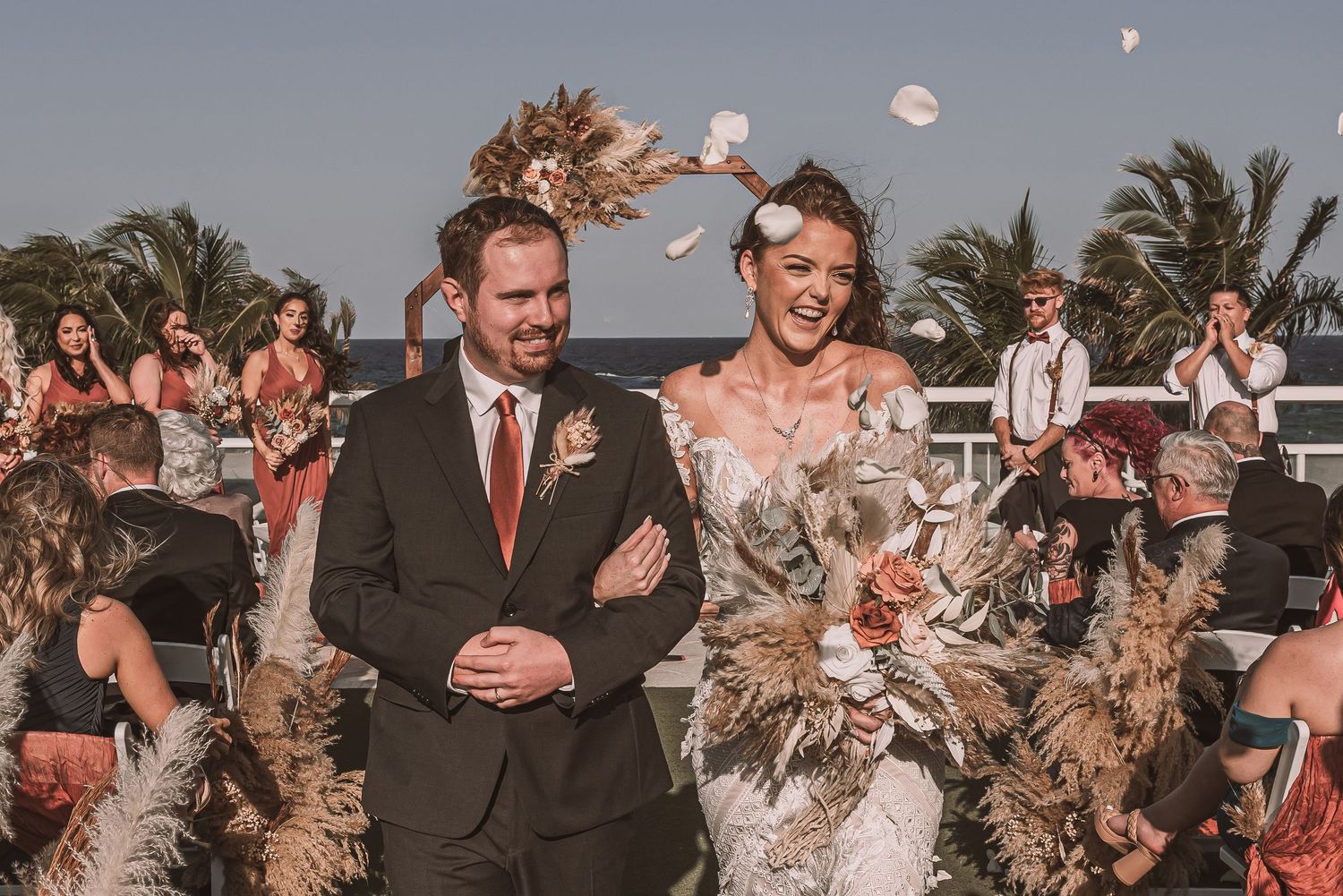 A wedding celebration during golden hour with guests tossing flower petals and palm trees in the background.