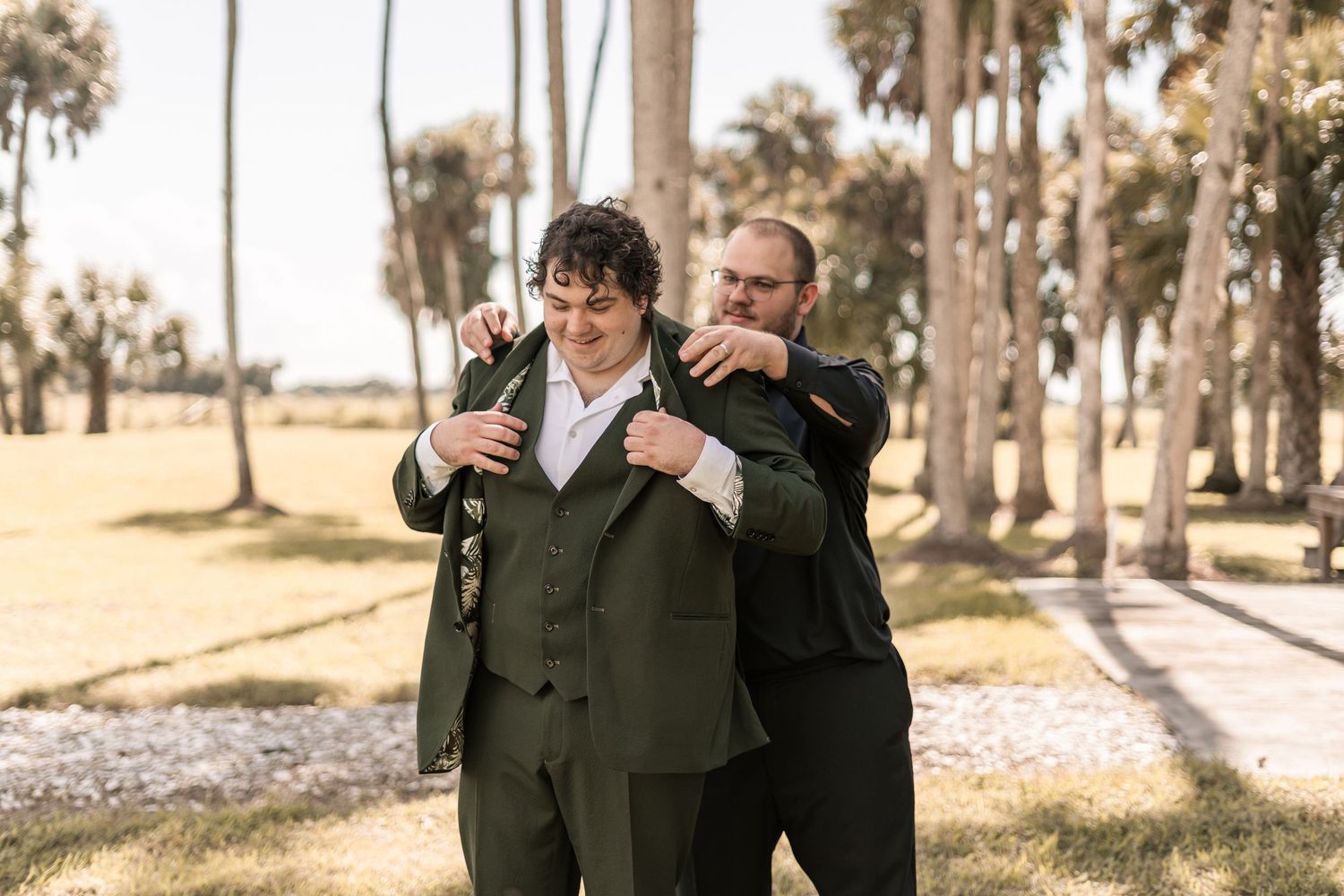 Two people in dark suits share a candid moment while standing among palm trees in an outdoor setting.
