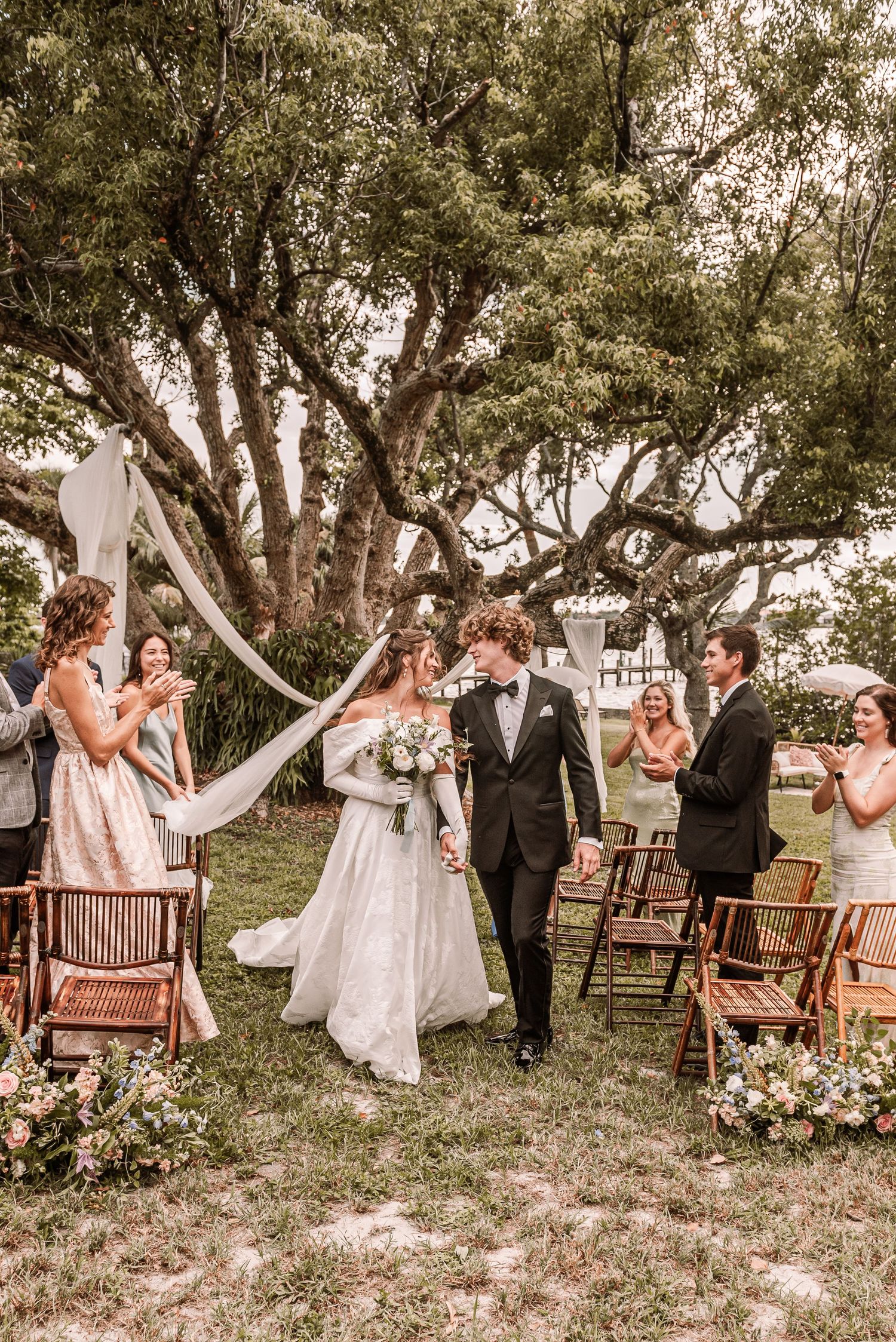 A romantic outdoor wedding ceremony under towering trees with white fabric draping and wooden chairs decorated with flowers.