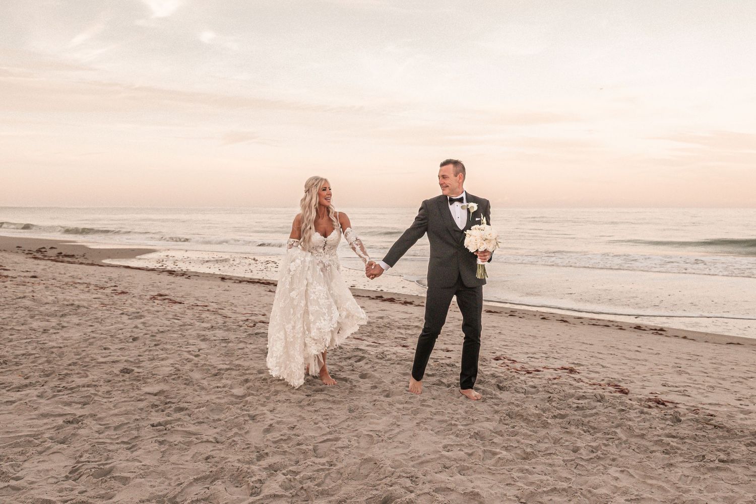 A couple in wedding attire runs joyfully along a sandy beach at sunset, with ocean waves in the background.