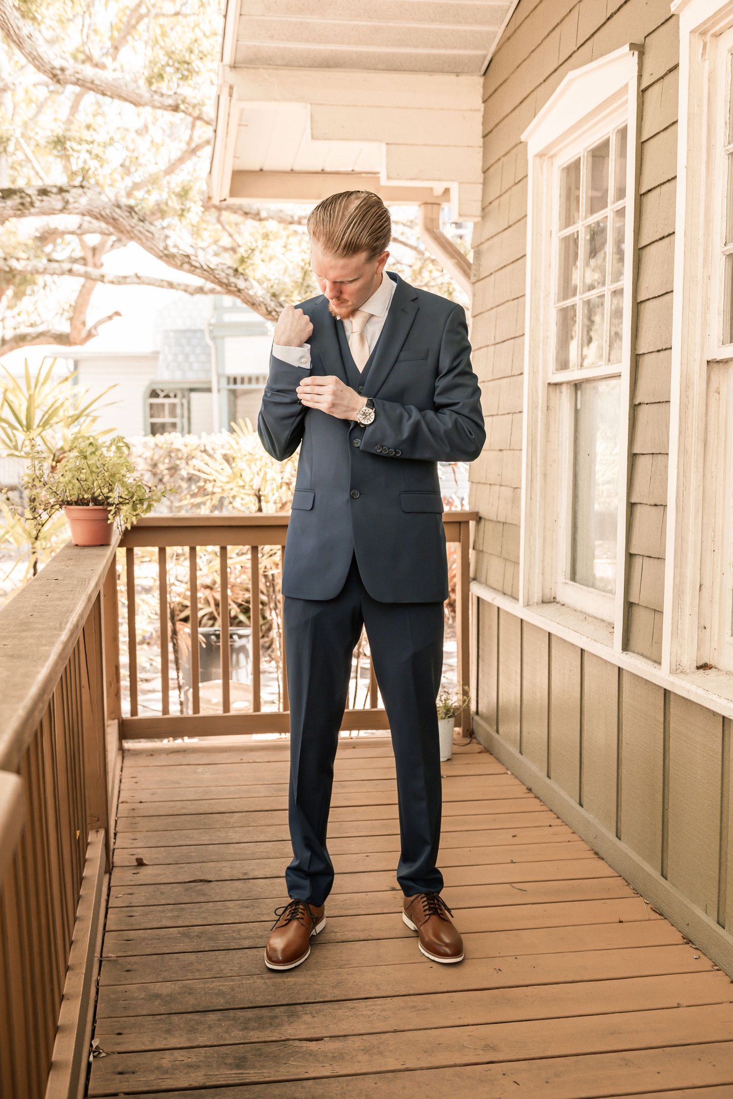 A groom in a navy suit adjusts his cufflinks on a wooden porch before his wedding ceremony.