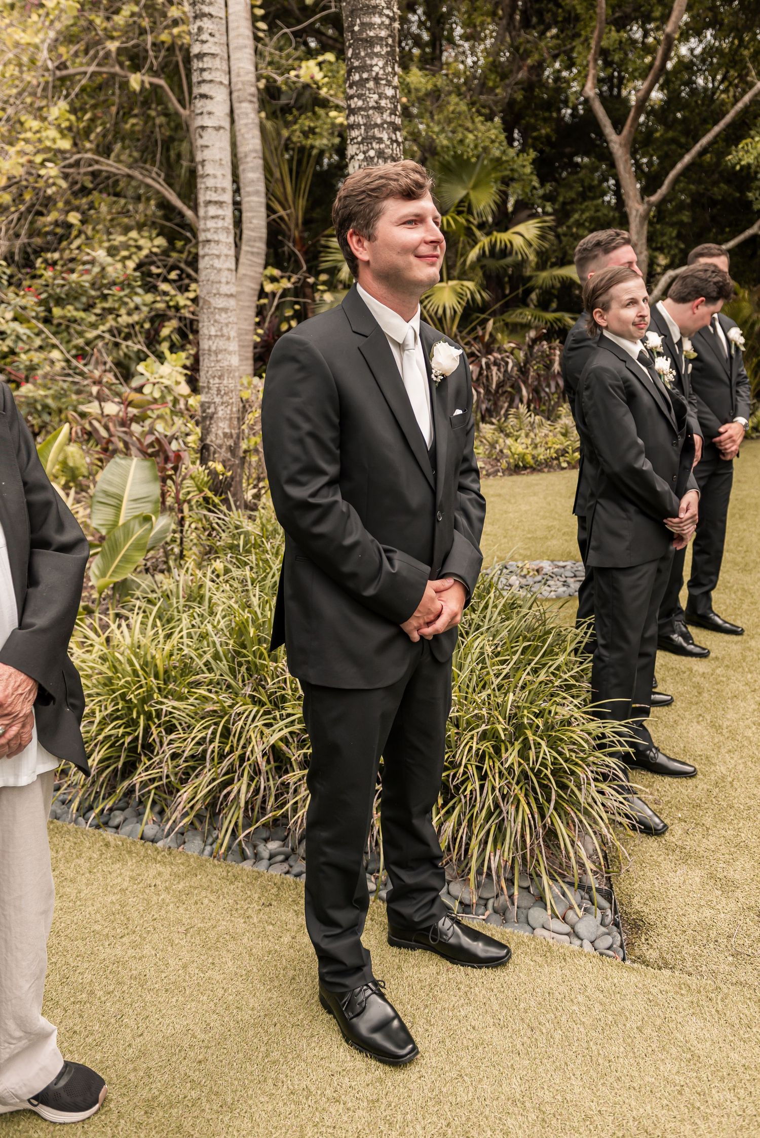 Groom in black tuxedo smiling at altar during outdoor garden wedding ceremony with groomsmen standing behind.