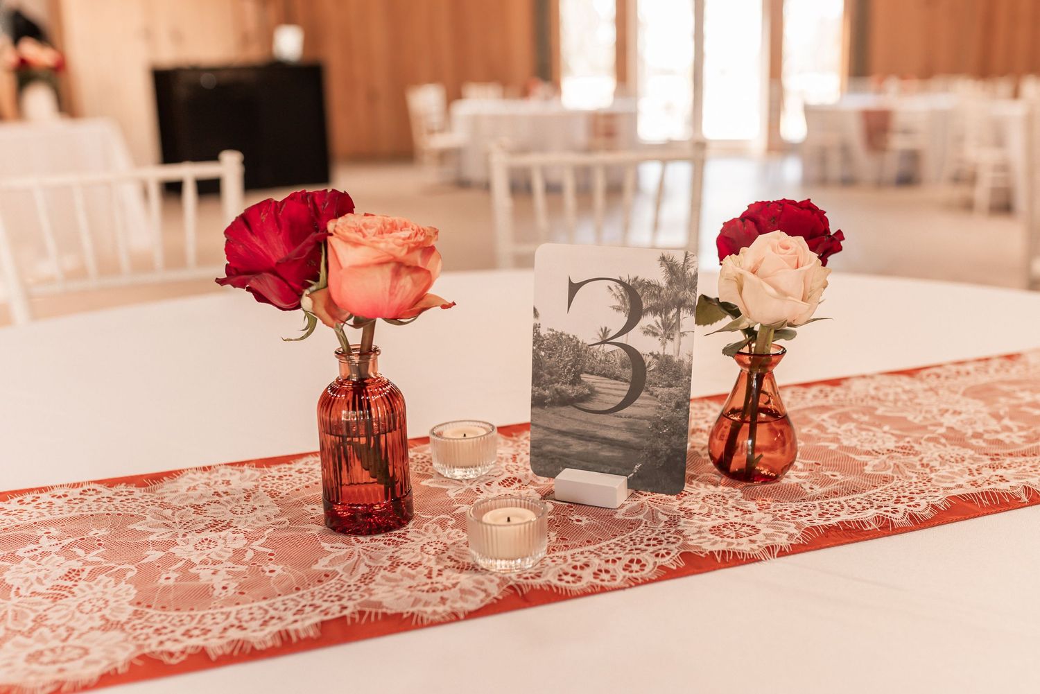 Red glass vases with floral arrangements line a table decorated with white linens and a red lace table runner.