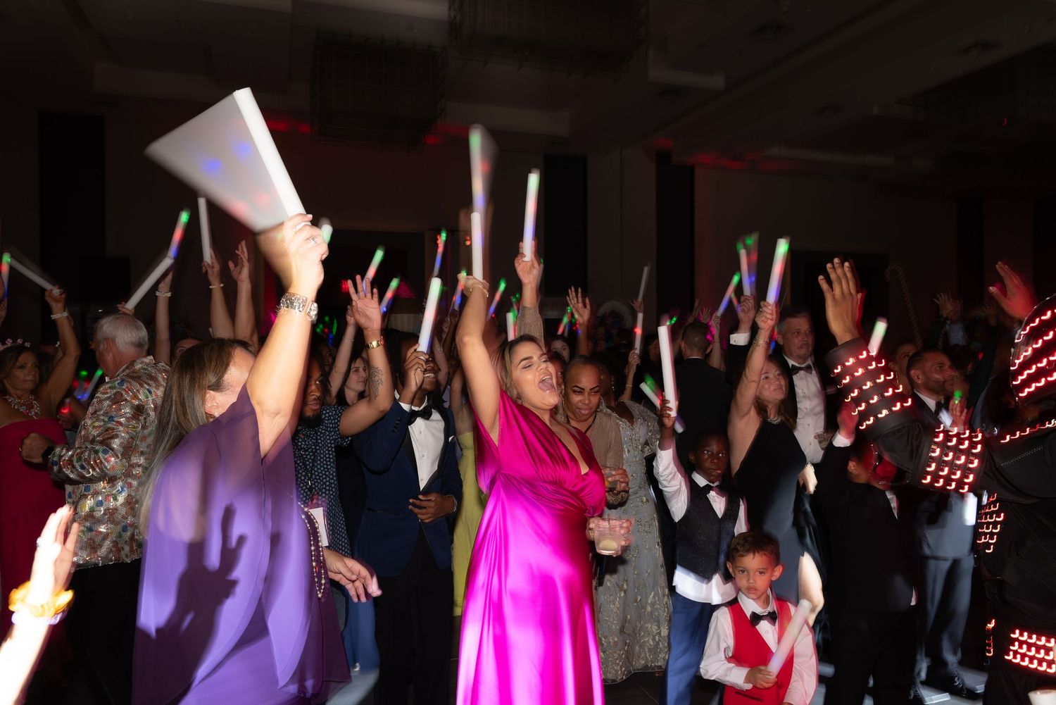 Crowd of people with raised arms dancing at a lively party with colorful lighting effects.