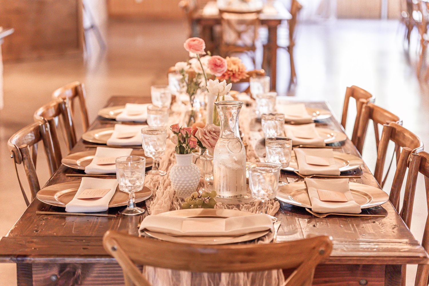 Elegant wedding reception tables set with white linens, wooden chairs, and soft floral centerpieces in a bright venue.