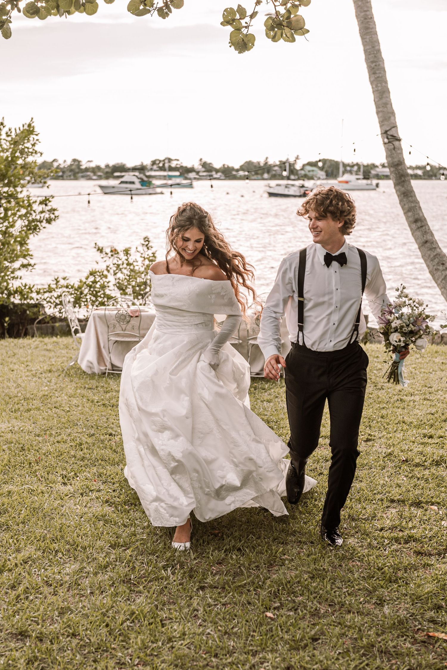 A couple in wedding attire playfully runs along a grassy waterfront area in a series of candid photos.
