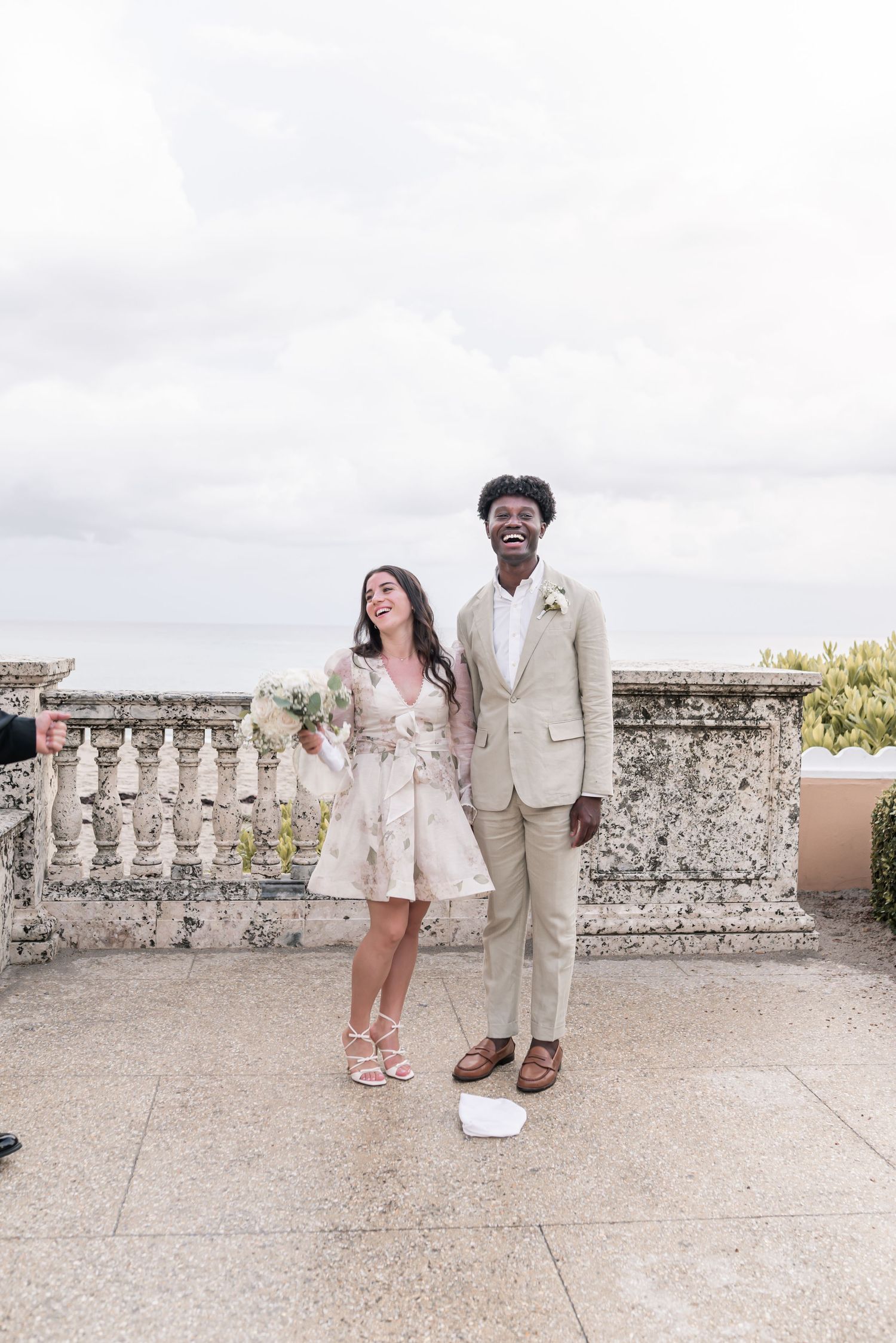 A couple in coordinated beige and floral outfits laughs together on a stone terrace during a photo session.