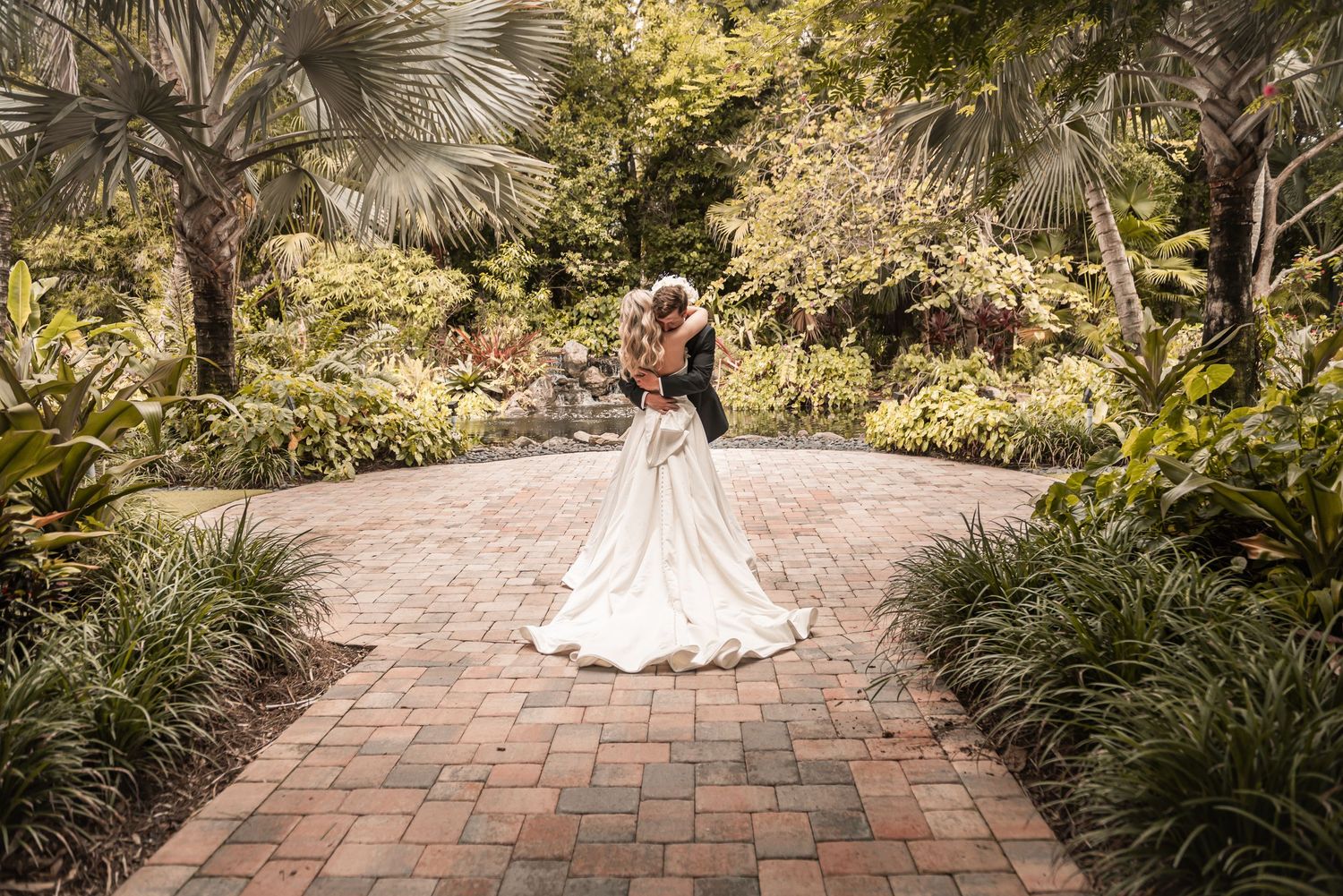 Bride and groom embracing on brick path surrounded by lush tropical garden greenery and palm trees.