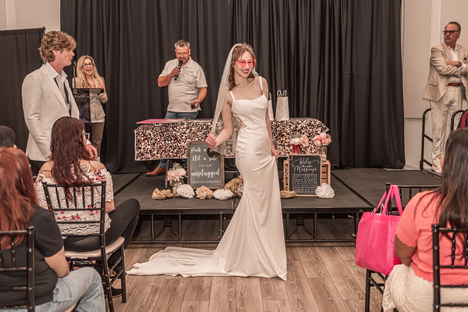 A bride stands in a fitted white mermaid wedding gown during a dress fitting with attendees and photographers nearby.