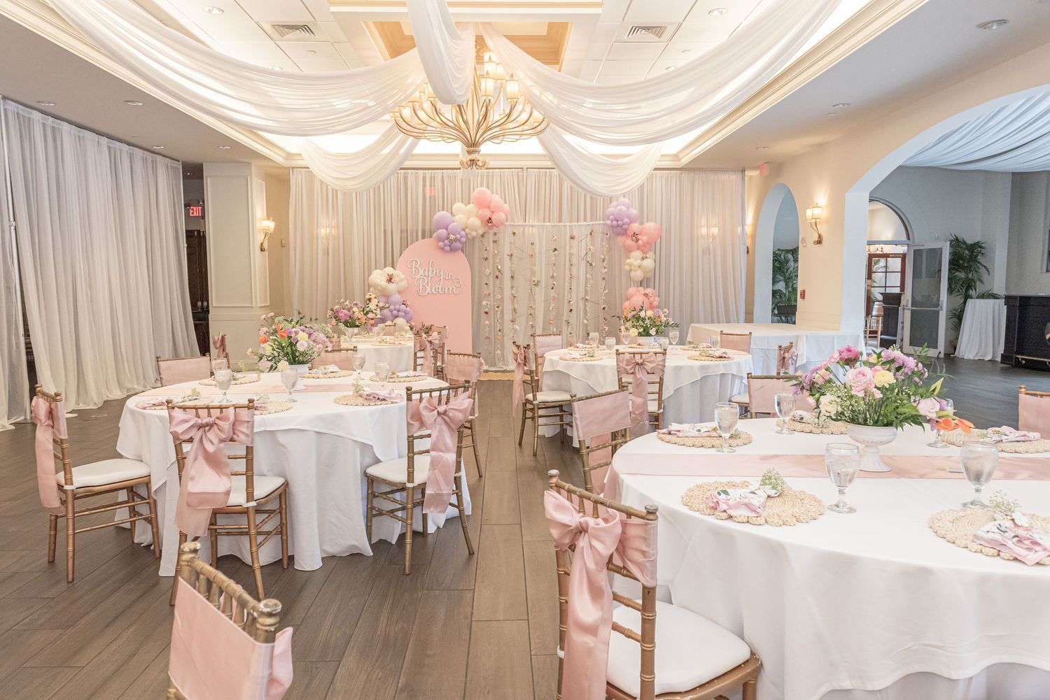 An elegant wedding reception room decorated in soft pink and white with round tables, floral centerpieces and draped fabric.