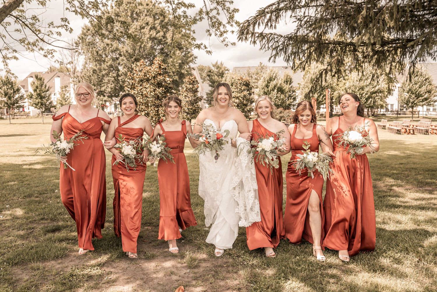 A wedding party wearing coral-colored bridesmaid dresses walks and laughs together outdoors in a sunlit garden setting.