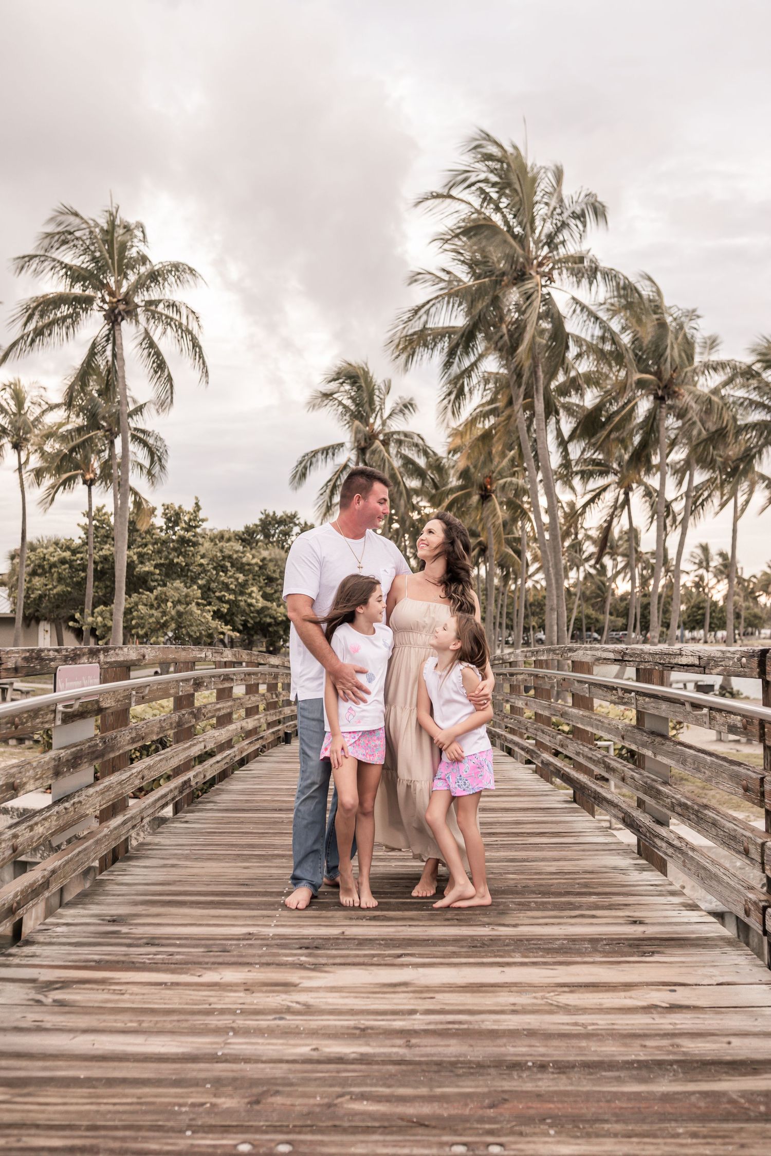 A family shares tender moments on a wooden boardwalk surrounded by swaying palm trees at sunset.
