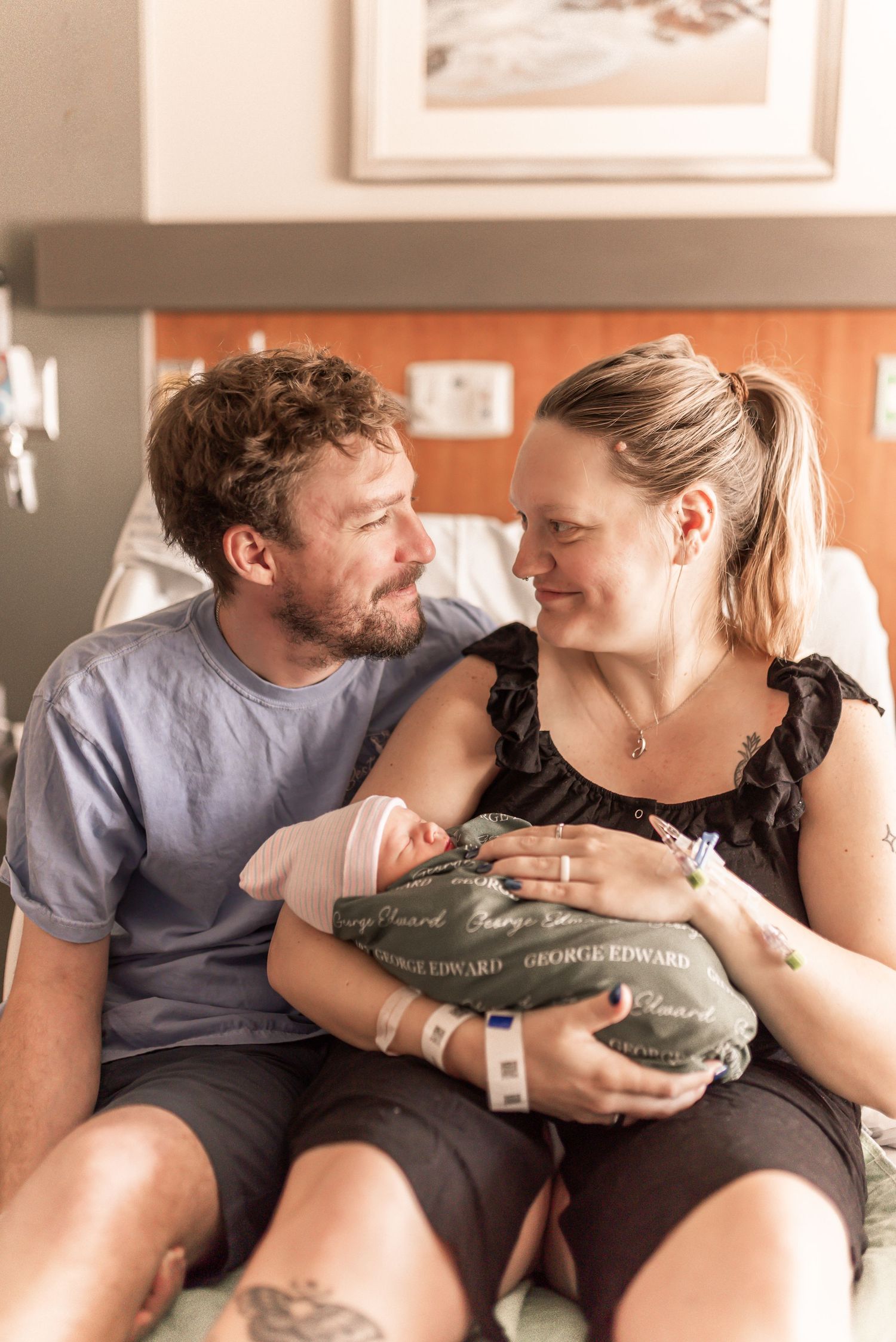 New parents smile at each other while holding their newborn baby wrapped in a green blanket in a hospital room.