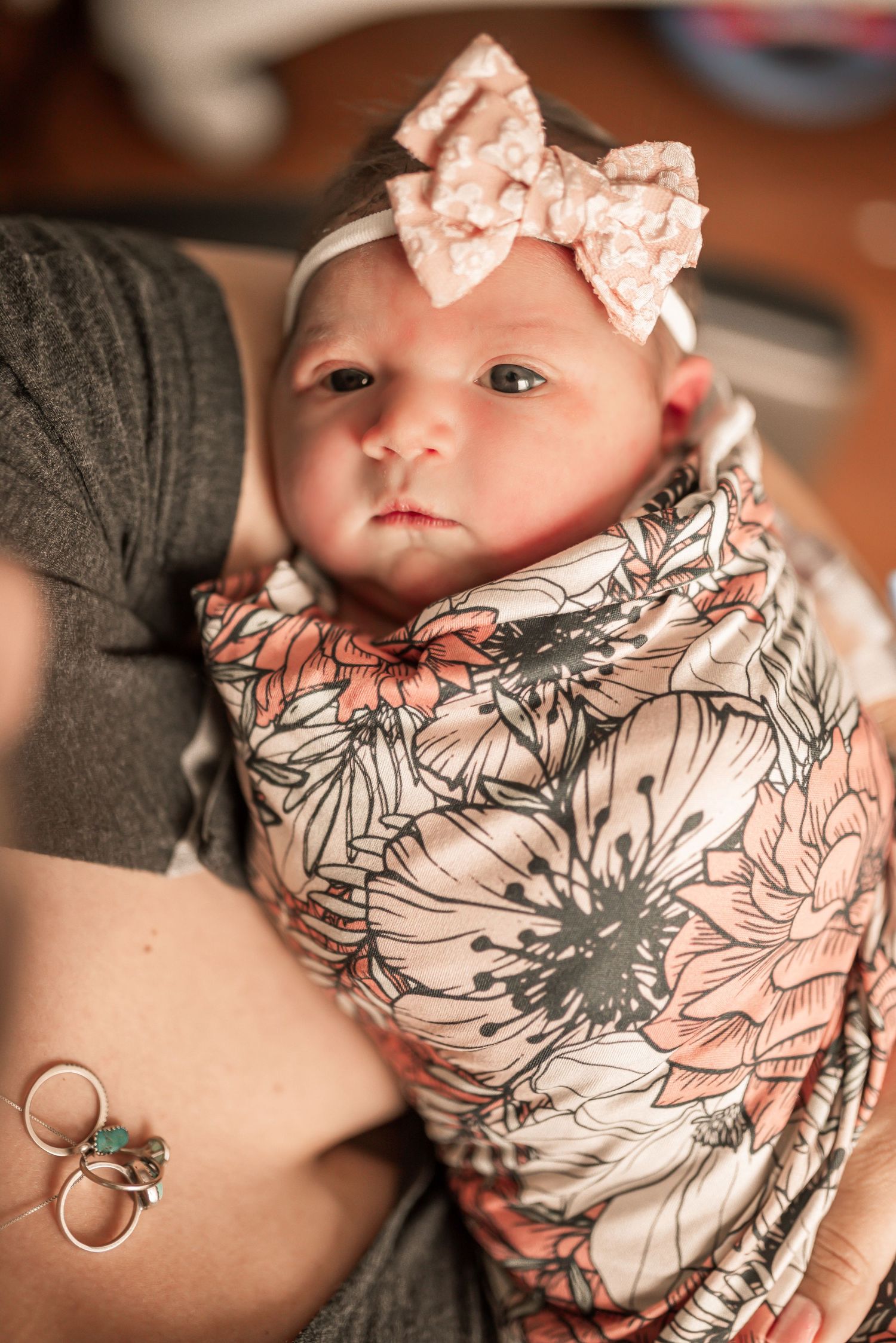 Newborn baby girl swaddled in floral blanket wearing pink bow headband, held by mother with rings visible.