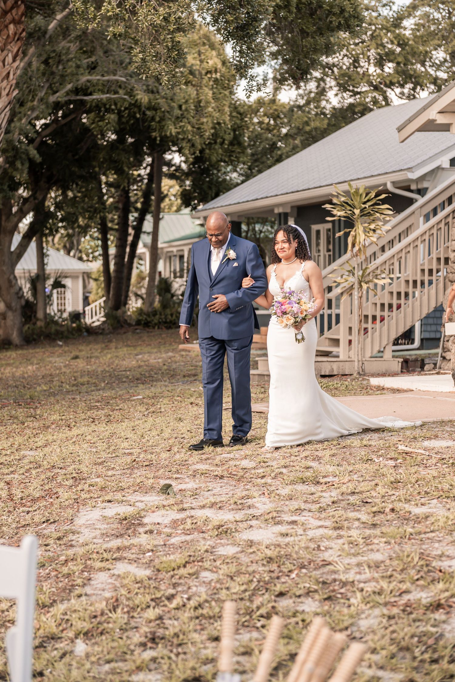 Bride in white gown walks down outdoor aisle with escort in blue suit, holding colorful bouquet at garden wedding.