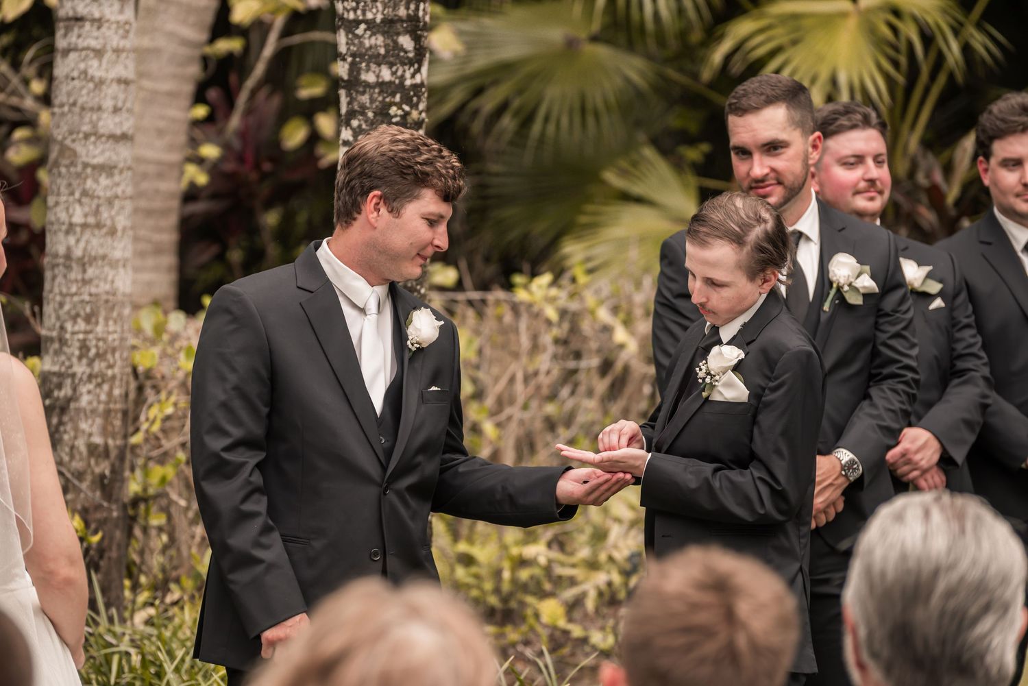 Groom receives wedding ring from young ring bearer during outdoor ceremony, with groomsmen standing in background.