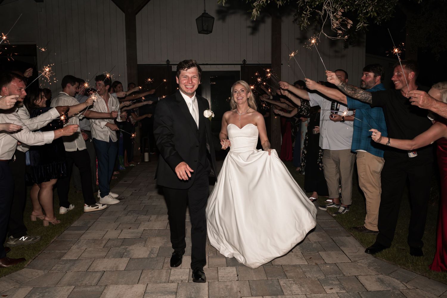 Bride and groom exit wedding reception through sparkler tunnel surrounded by cheering guests at night.
