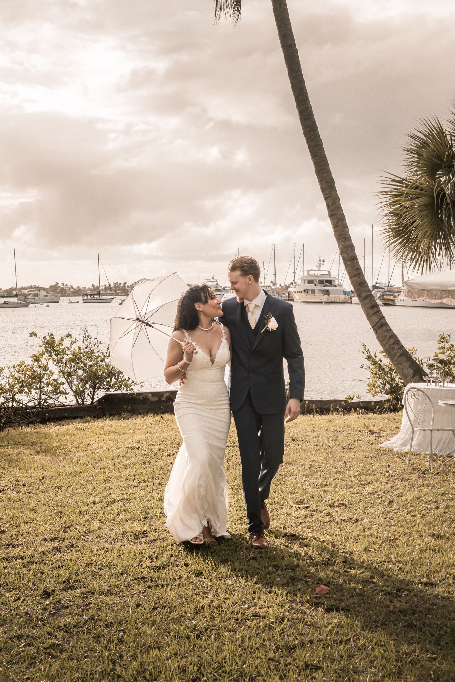 Bride and groom walk on tropical waterfront lawn, she holds clear umbrella, boats visible behind them.