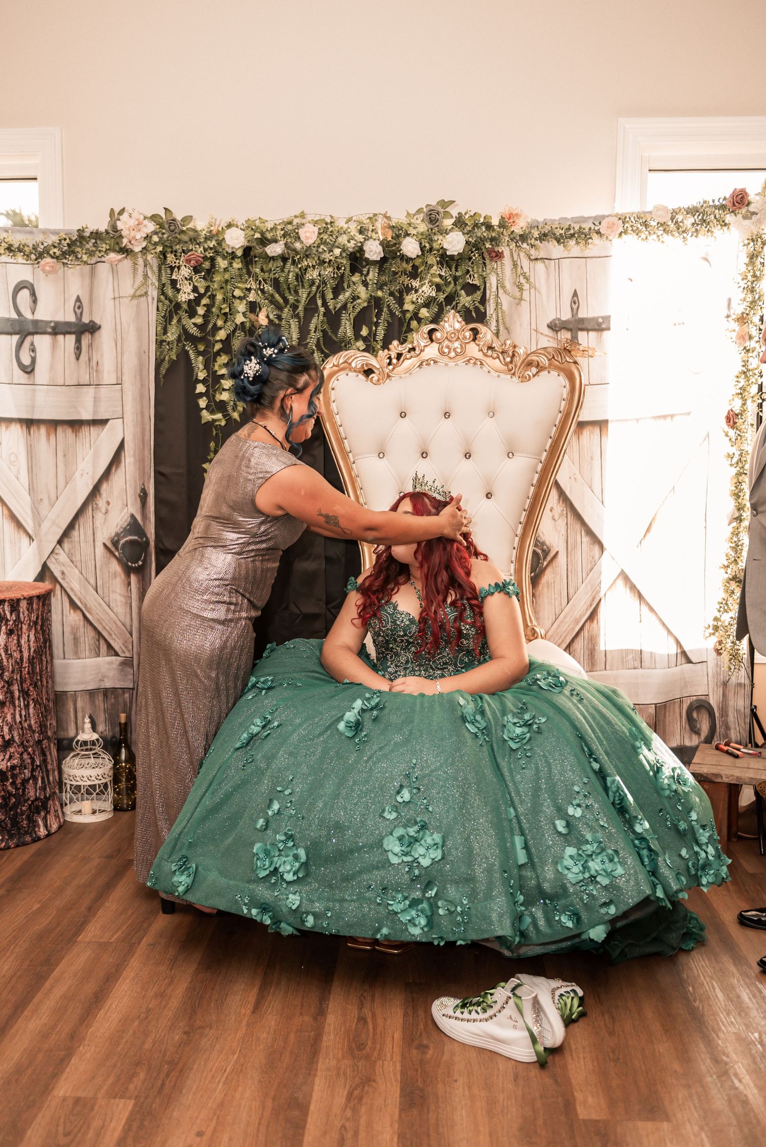 A young woman in a green quinceañera gown sits on a throne chair as someone places a tiara on her head.