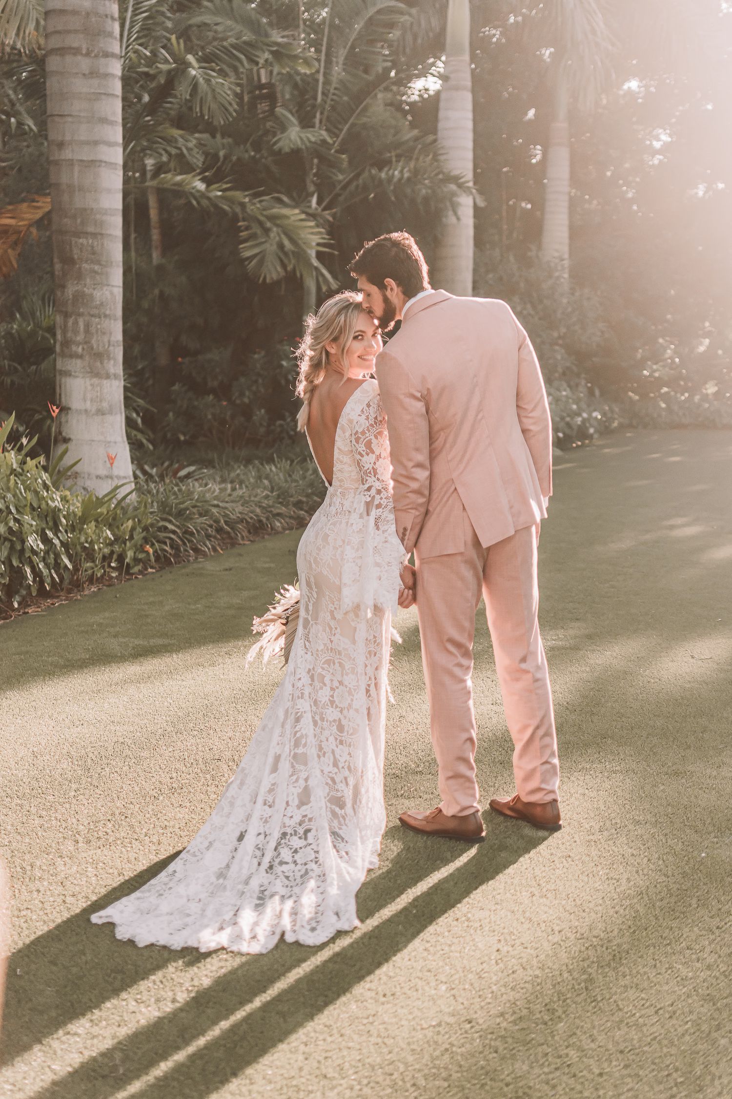 A romantic wedding photo of a couple in pink suit and lace dress standing among palm trees in soft sunlight.