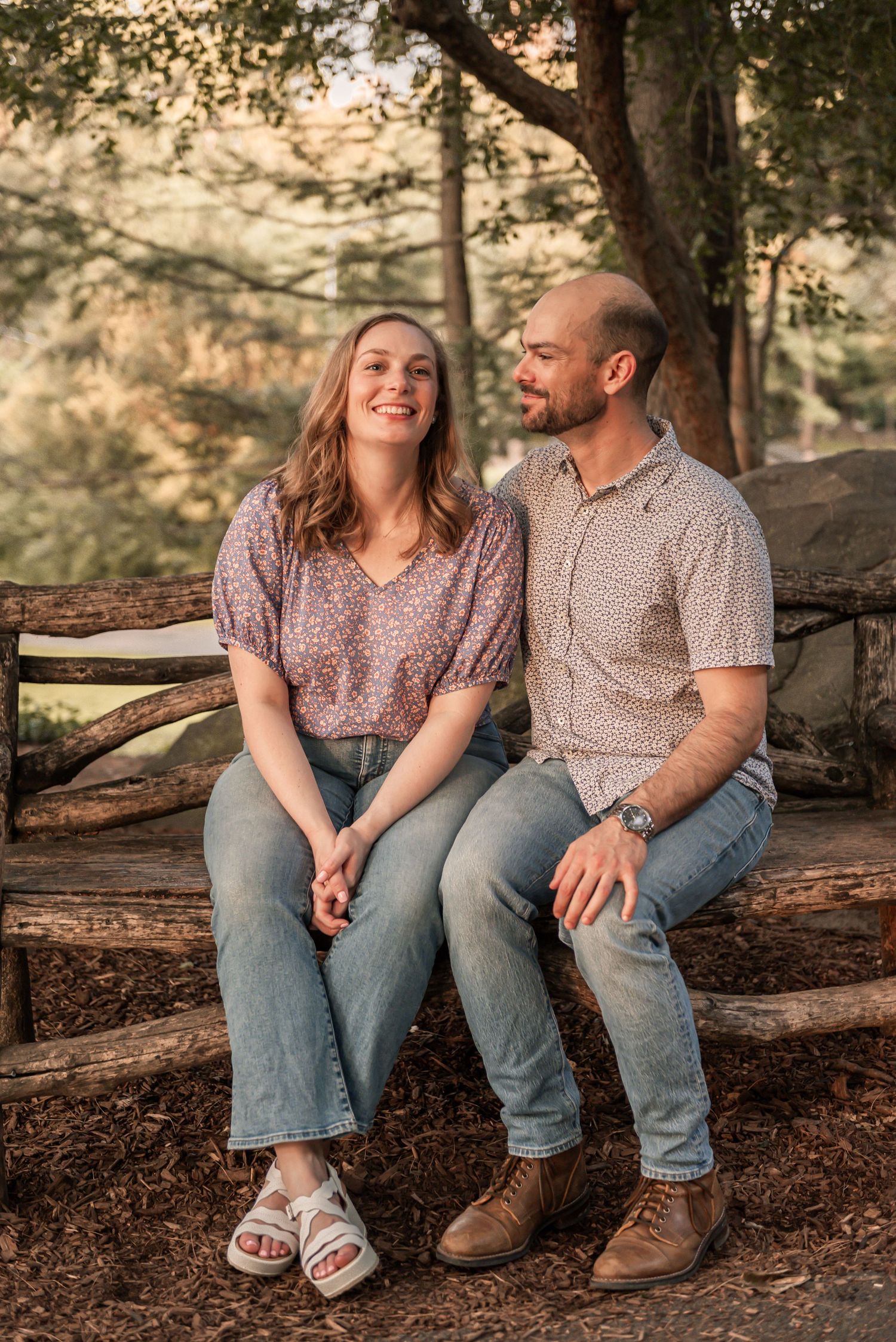A couple shares a joyful moment while sitting together on a rustic wooden fence in a wooded setting at sunset.