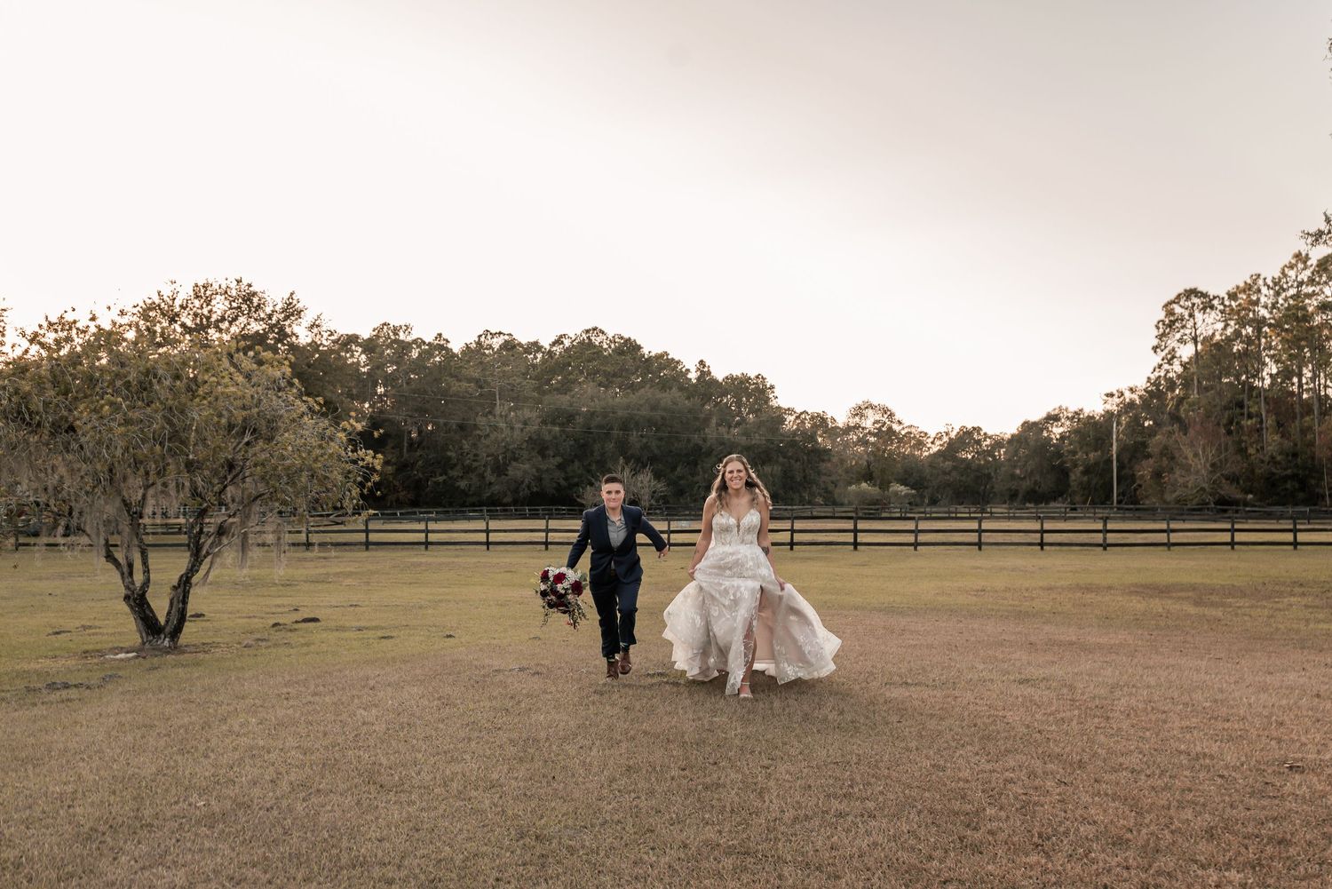 A couple in wedding attire walks through a rustic field at sunset with trees in the background.