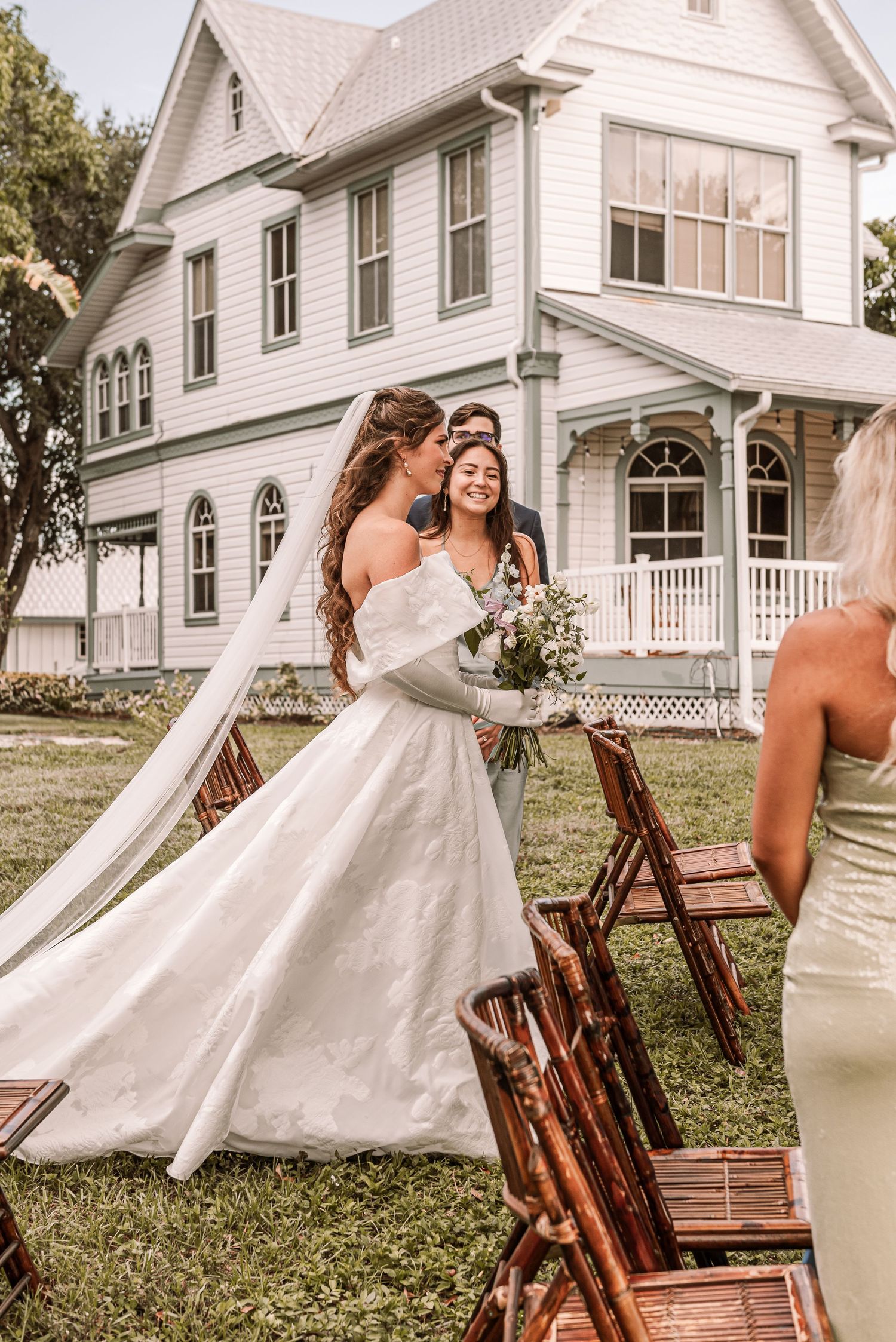 A joyful wedding moment captured outside a white Victorian-style house with wooden chairs in the foreground.