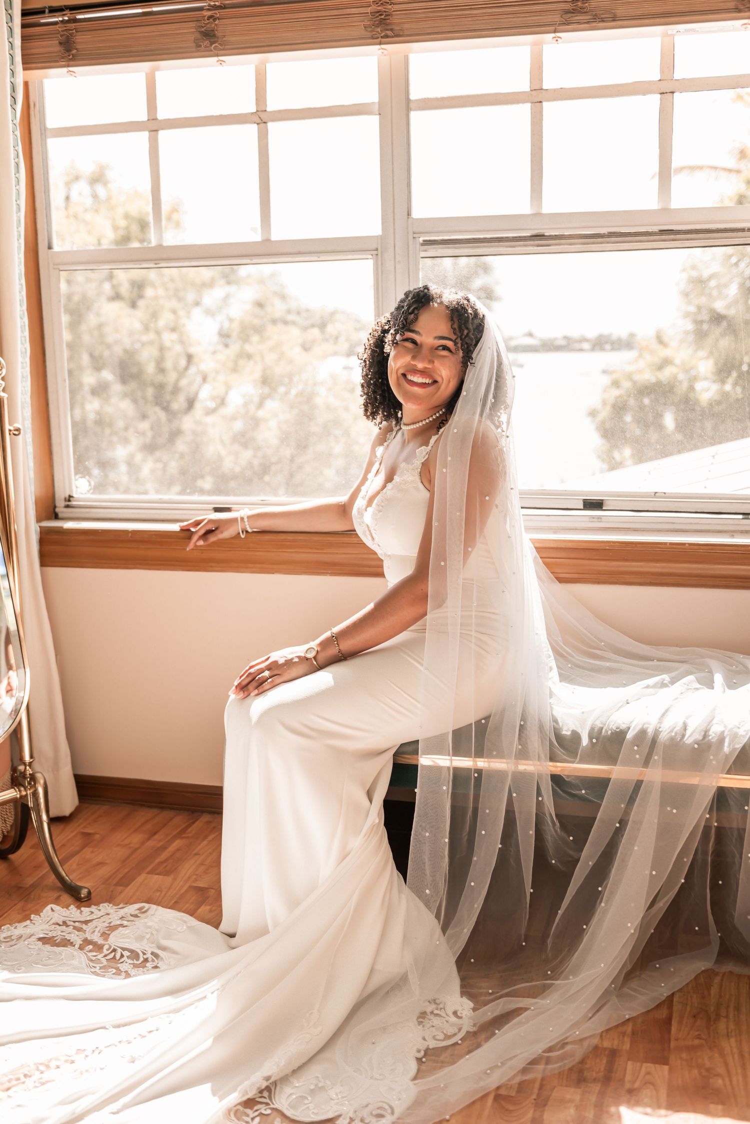 Smiling bride in white gown and long veil sits by sunlit window on her wedding day.