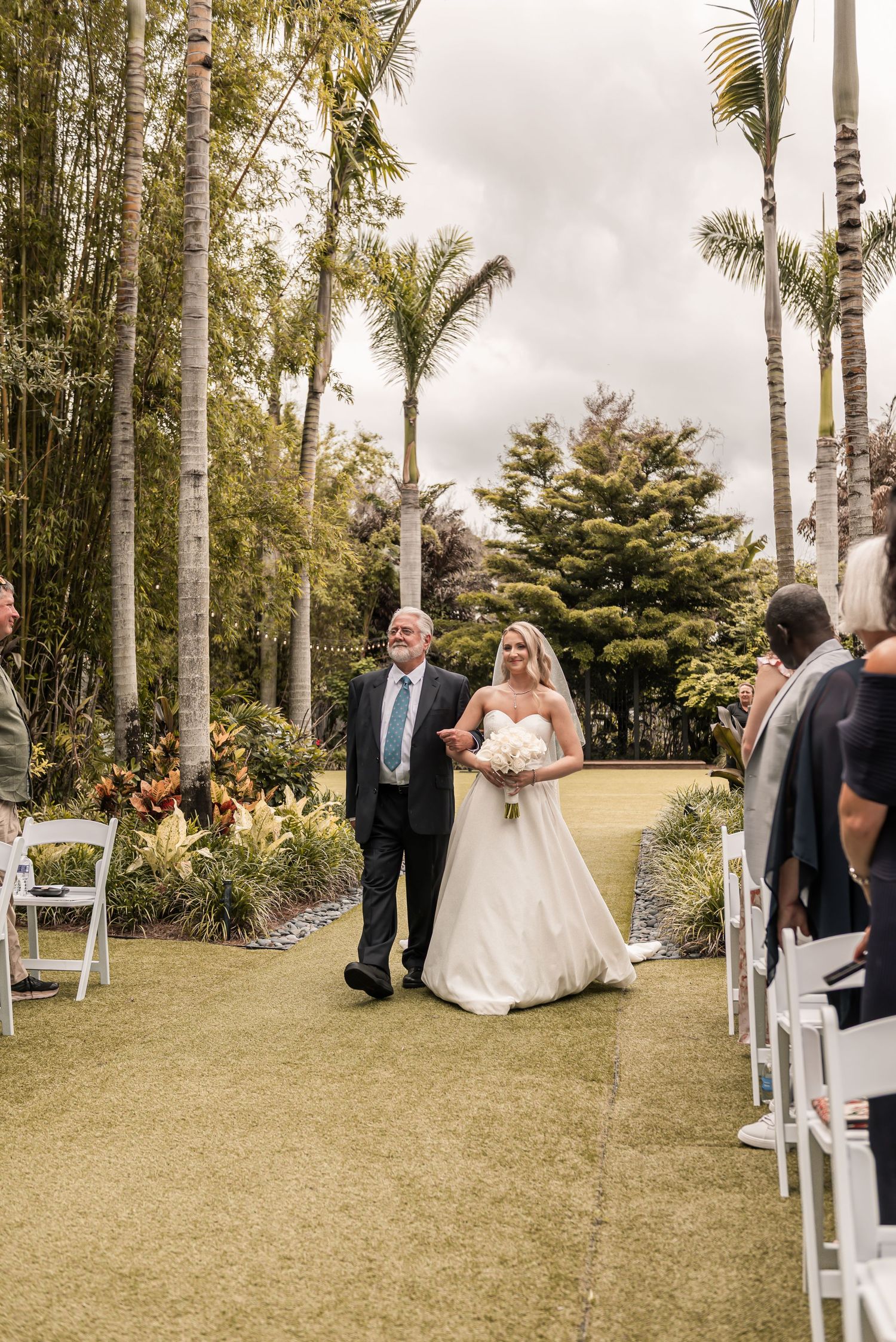Bride walks down outdoor aisle with escort among palm trees at garden wedding ceremony.