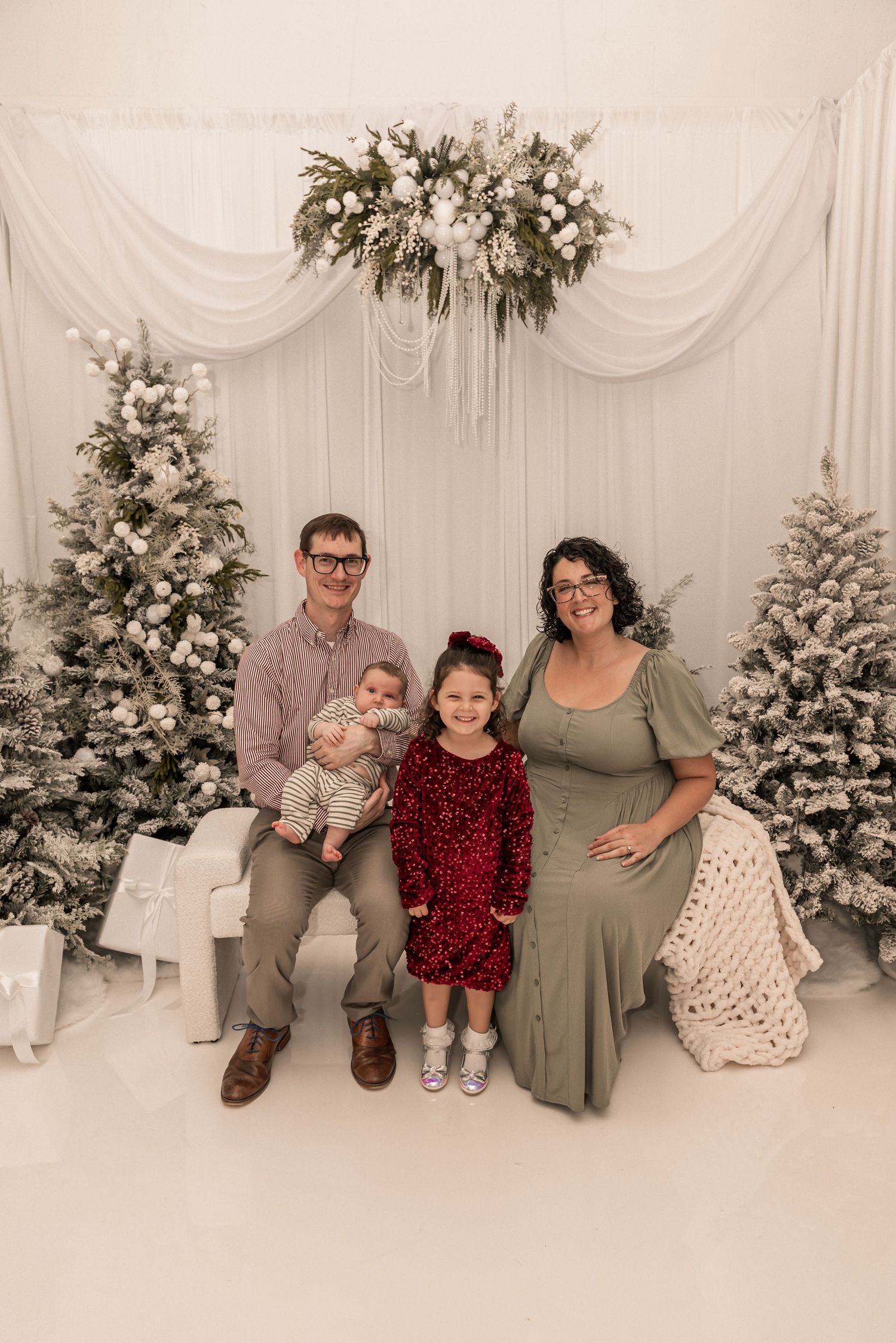 A family poses in front of decorated Christmas trees and floral arrangements in a white studio setting.