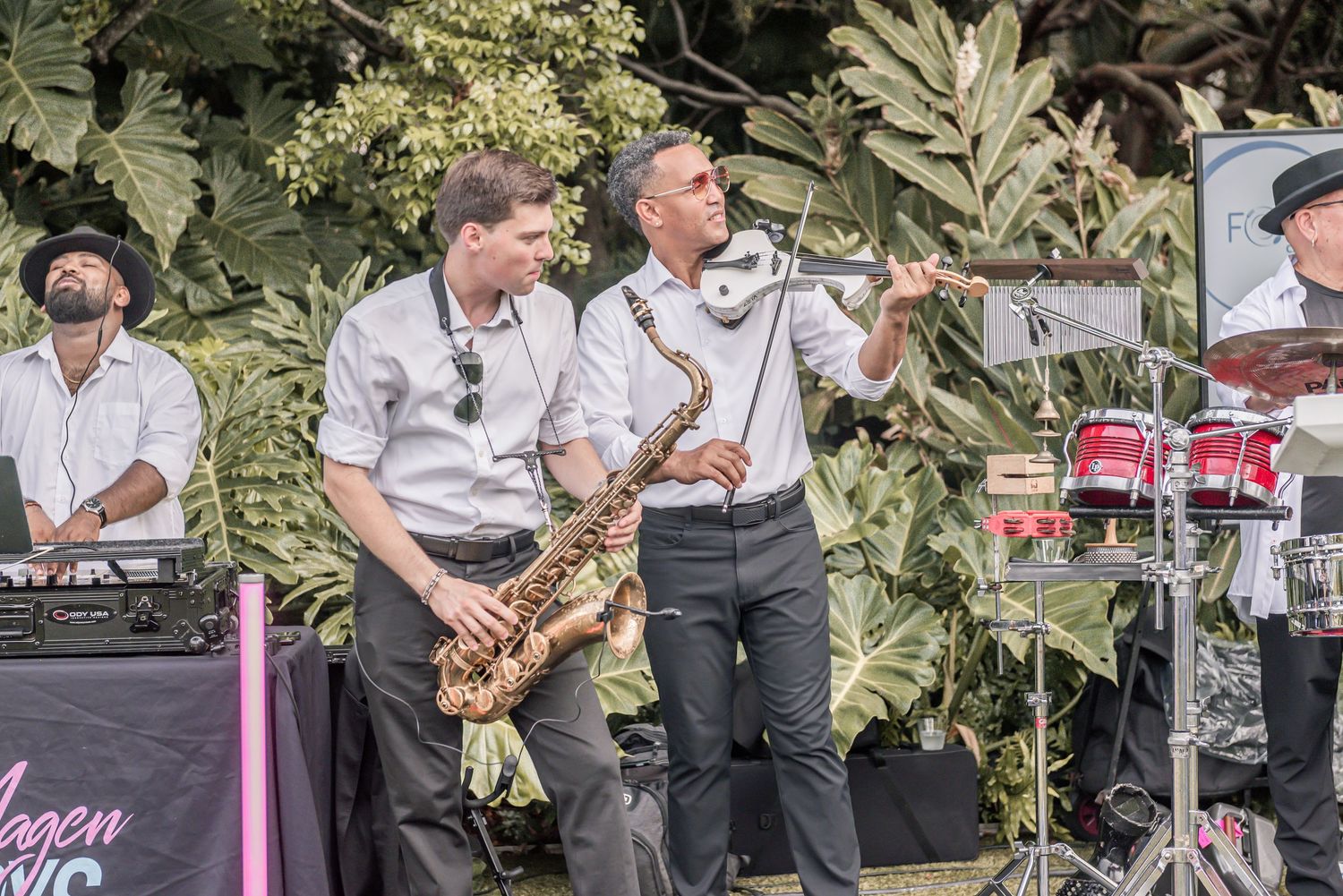 Musicians perform on outdoor stage with saxophone and vocalist backed by tropical foliage and drum kit.