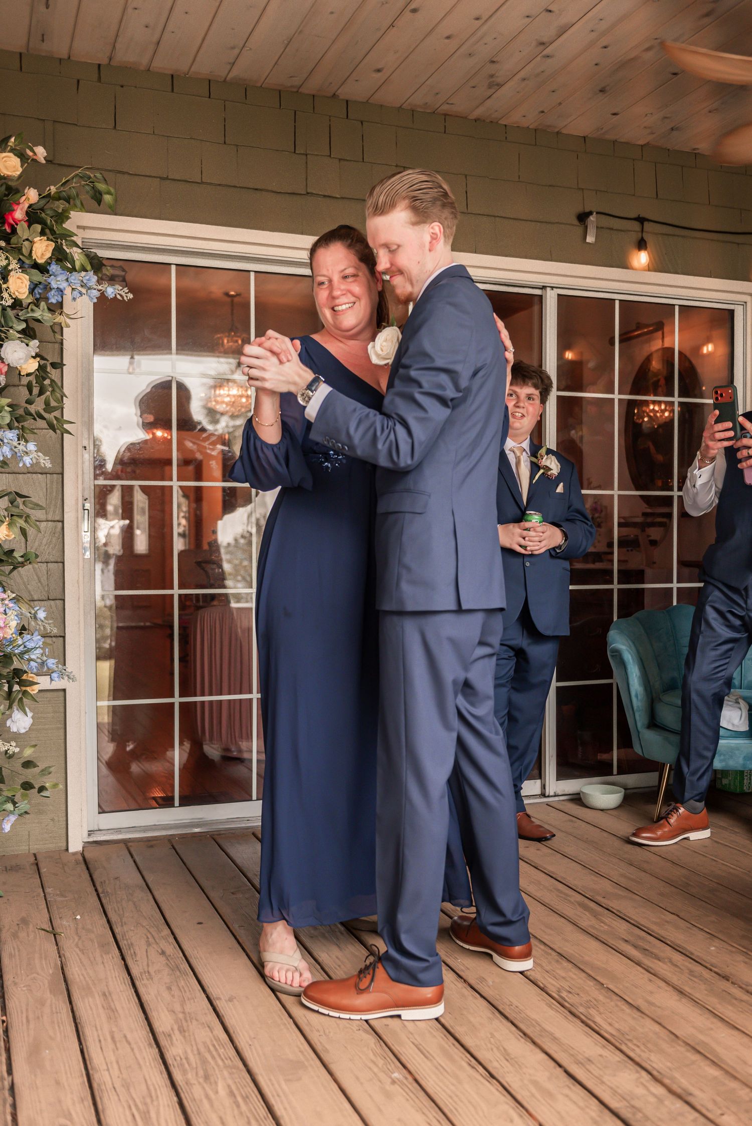 Mother and son share a heartfelt dance on a wooden deck during a wedding reception, both dressed in navy blue.