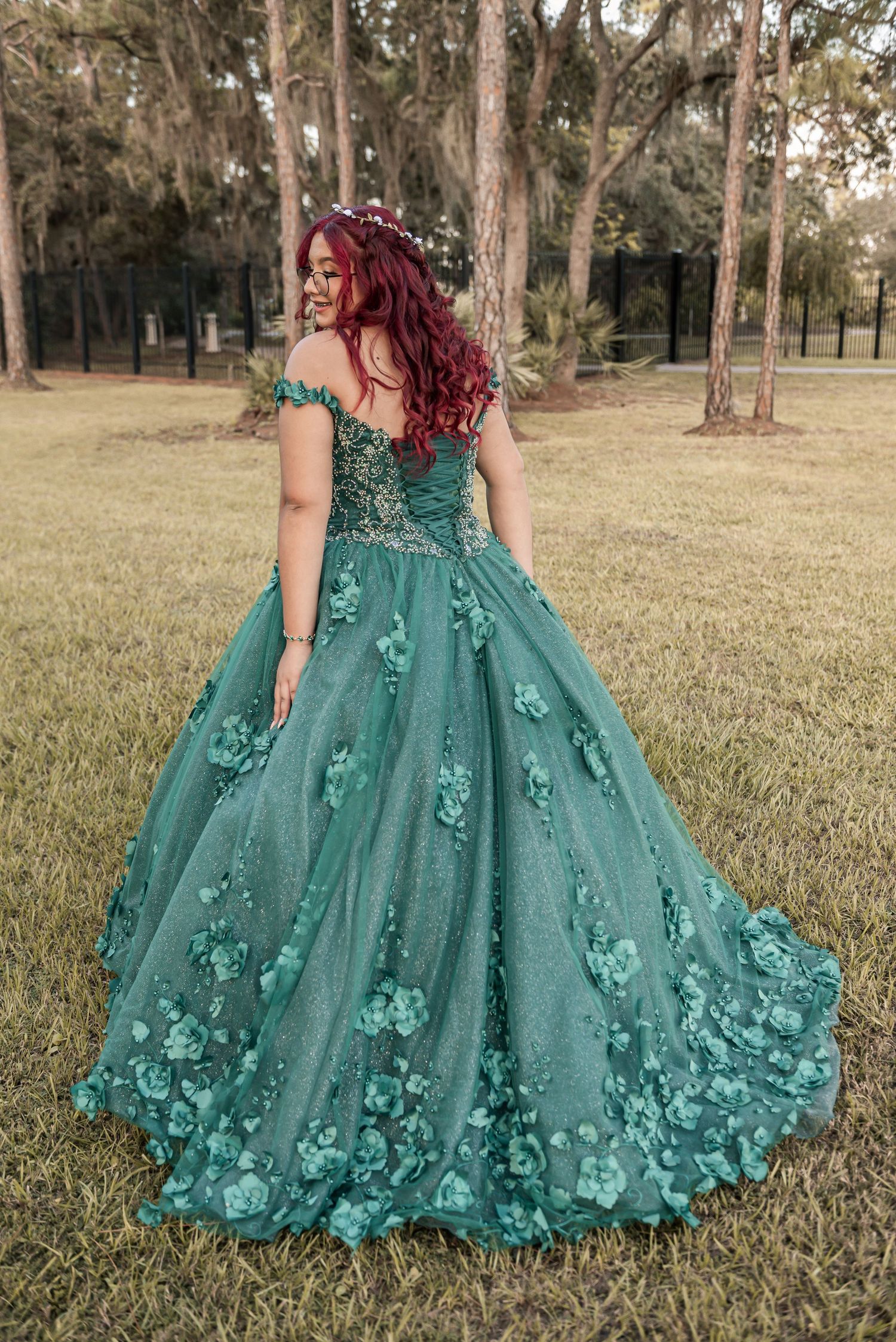 Young woman in elegant teal green off-shoulder ball gown with 3D floral appliques posing outdoors in a garden setting.