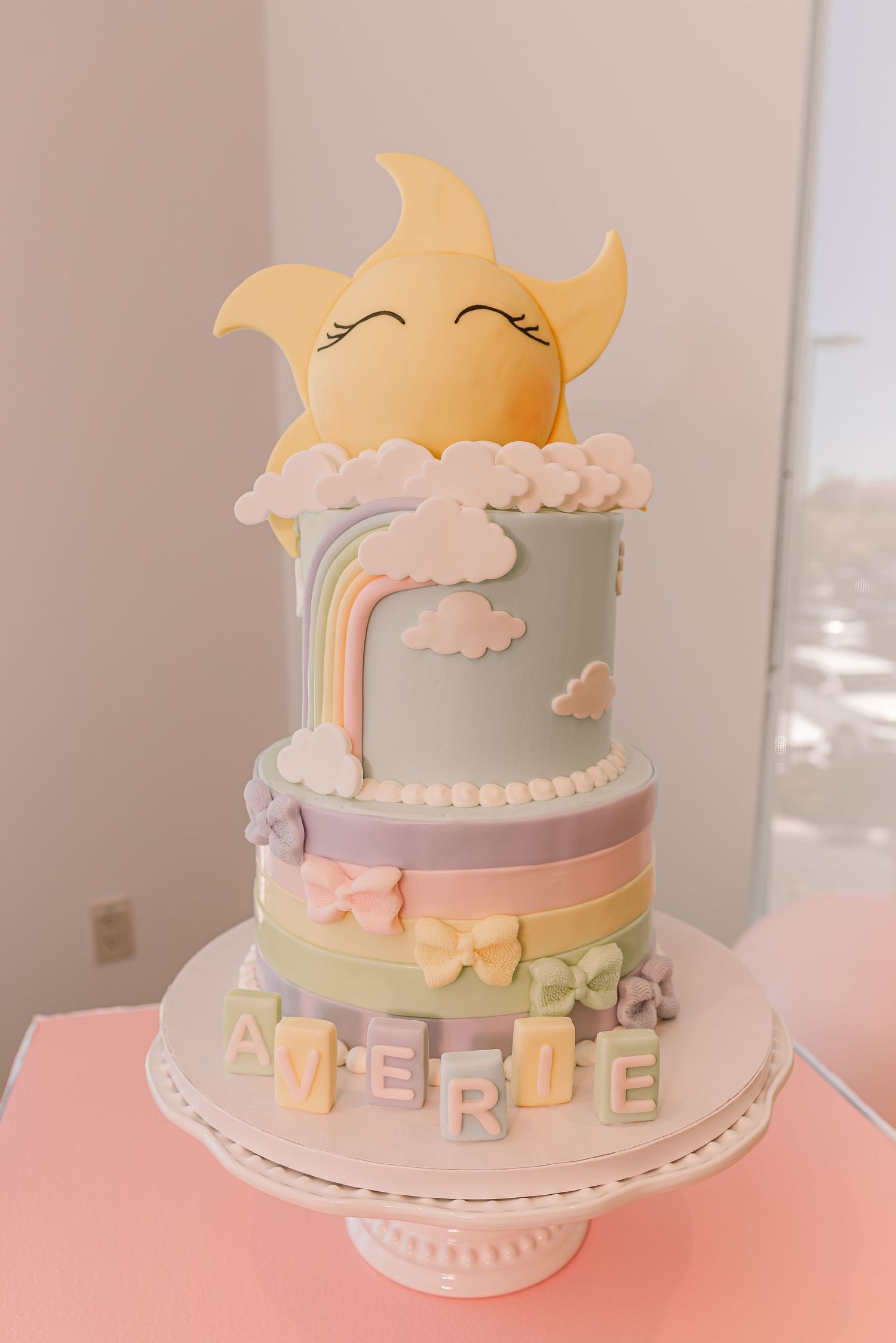 Two-tiered pastel rainbow cake with smiling sun topper, soft clouds, and name letters in pink and yellow tones.