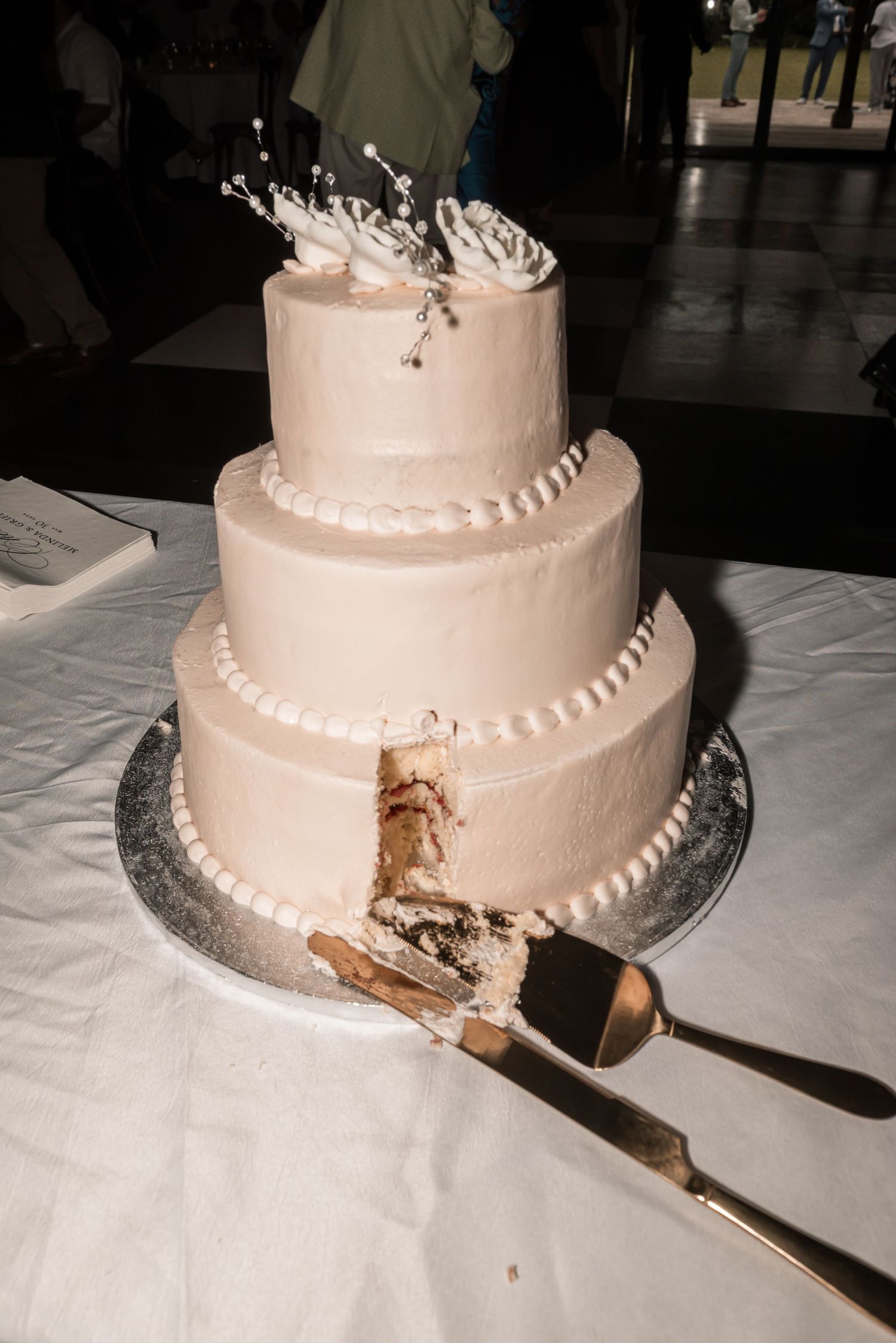 Three-tier white wedding cake with floral topper, partially sliced, displayed on silver board with cake server.