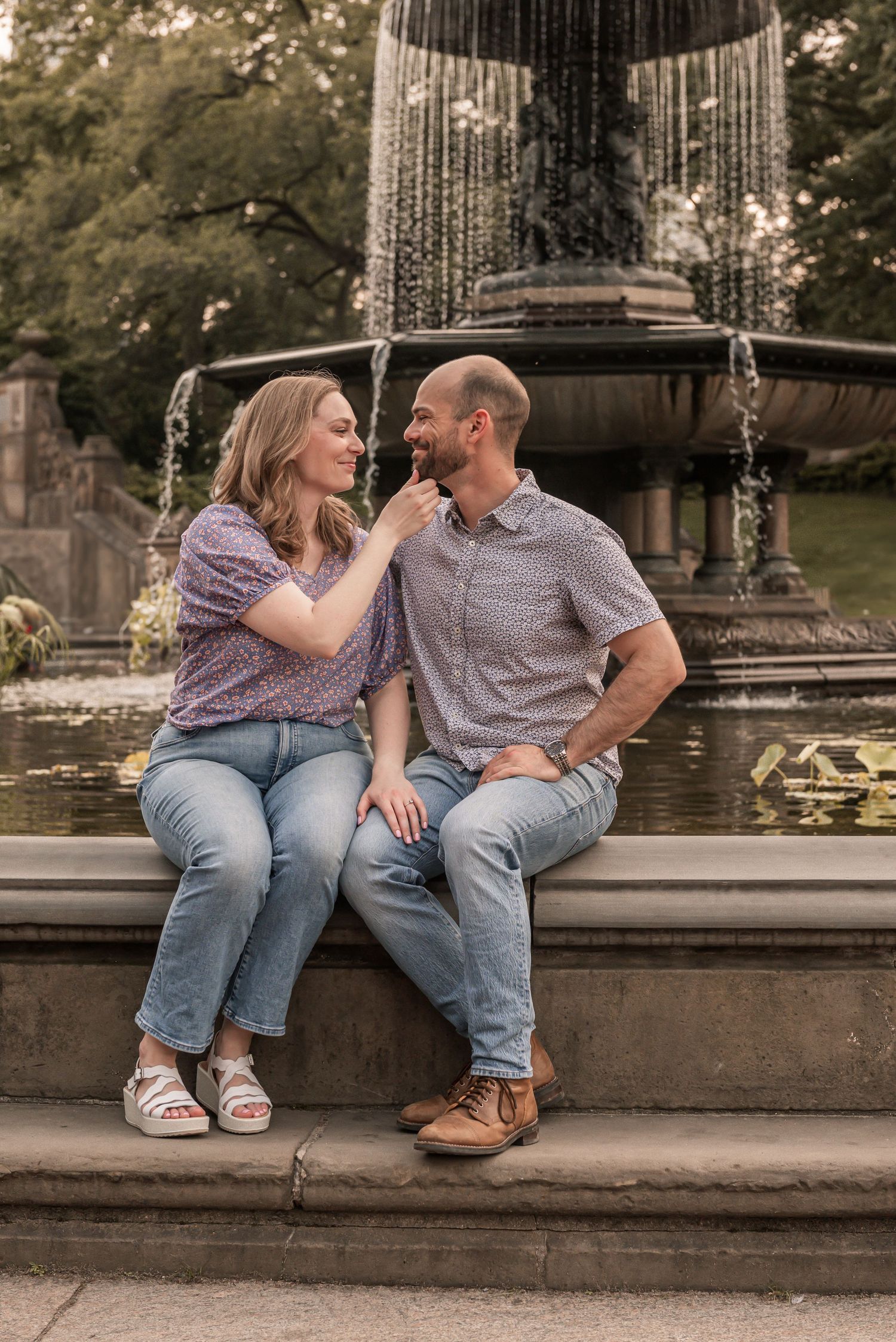 A couple sits together on stone fountain steps in a romantic setting during an engagement photo session at a park.