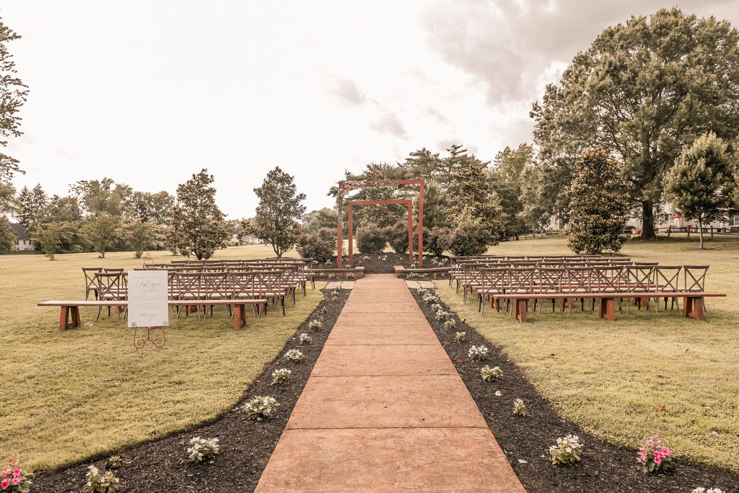Long brick pathway in an outdoor garden leads to rustic barn structures and wooden benches under mature trees.