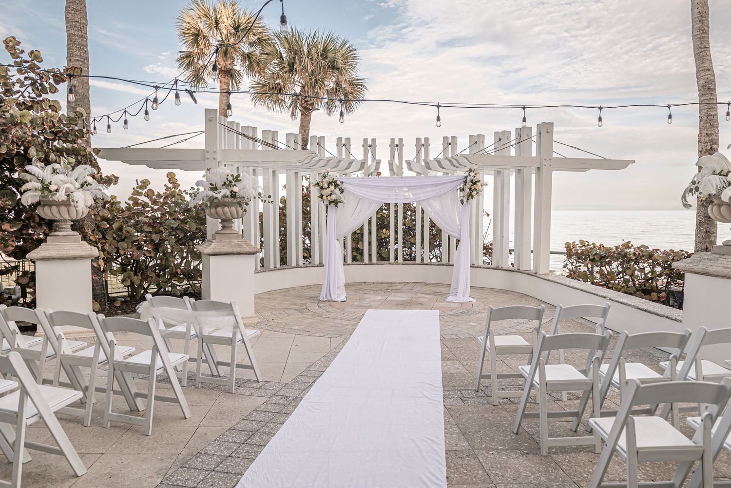 White folding chairs arranged for an outdoor beach wedding ceremony with palm trees and string lights overhead.