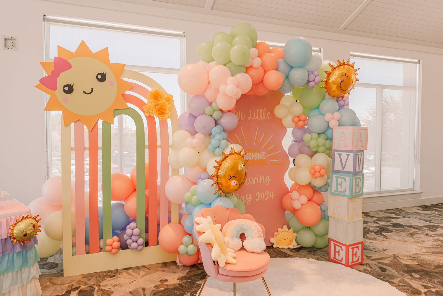 Pink and pastel balloon arch with smiling sun decoration and children's chair for birthday party photoshoot setup.