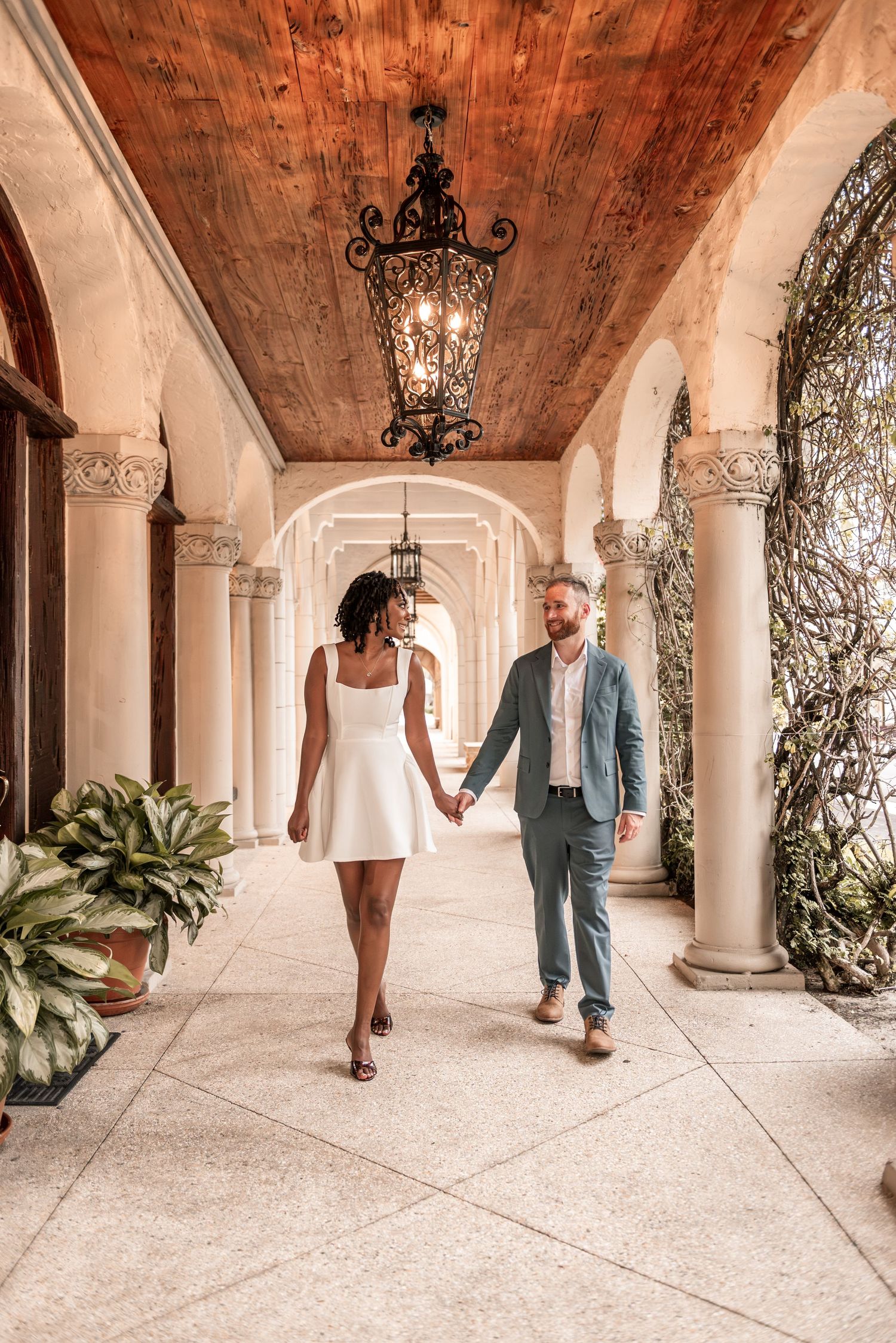 Couple holding hands walking through elegant arched colonnade with wood ceiling and hanging lantern.