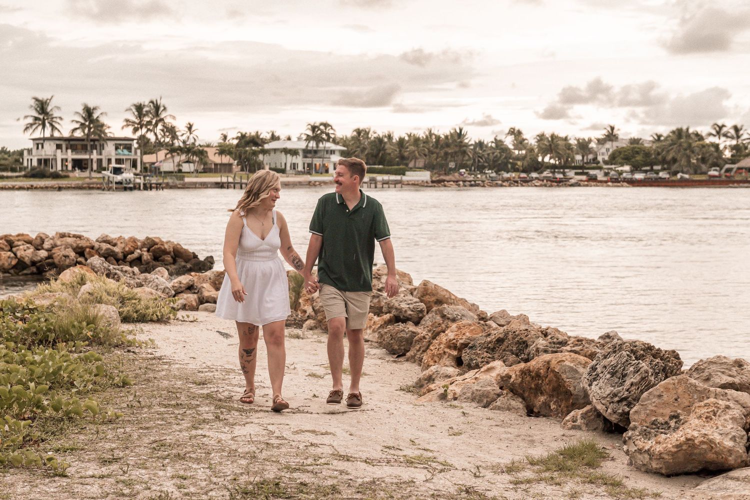 Couple walking hand in hand along a tropical beach with palm trees and resort buildings in the background.