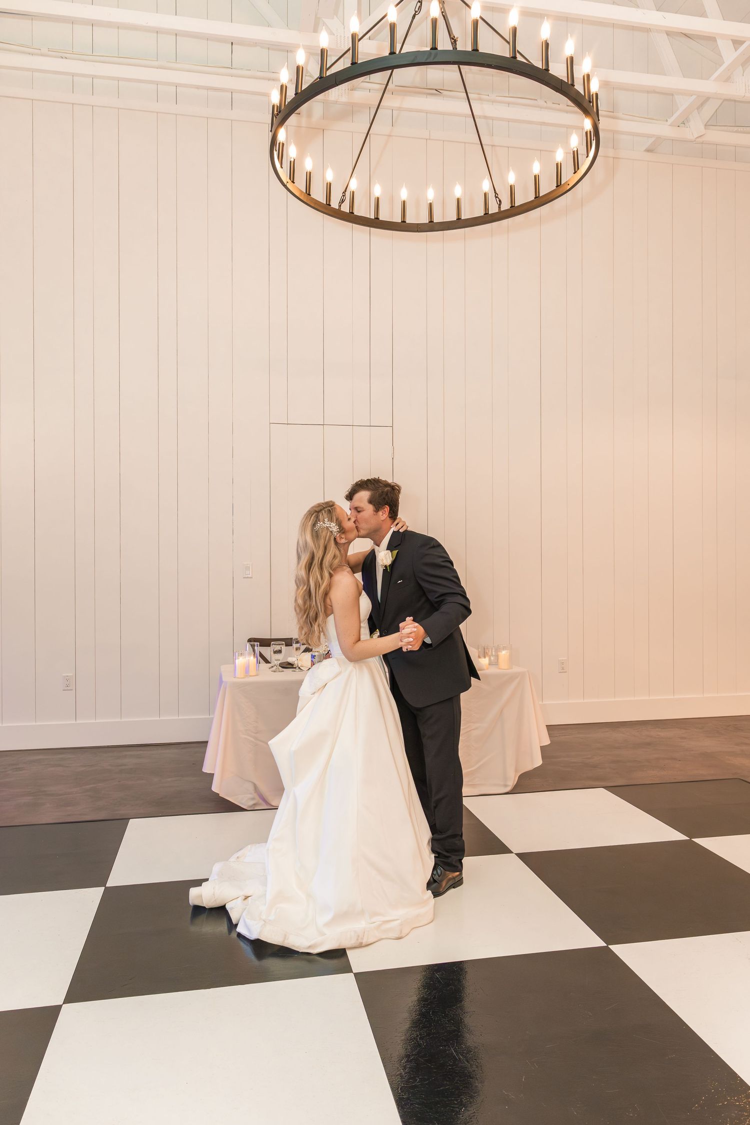 Bride and groom share first dance on black and white checkered floor beneath elegant circular candle chandelier.